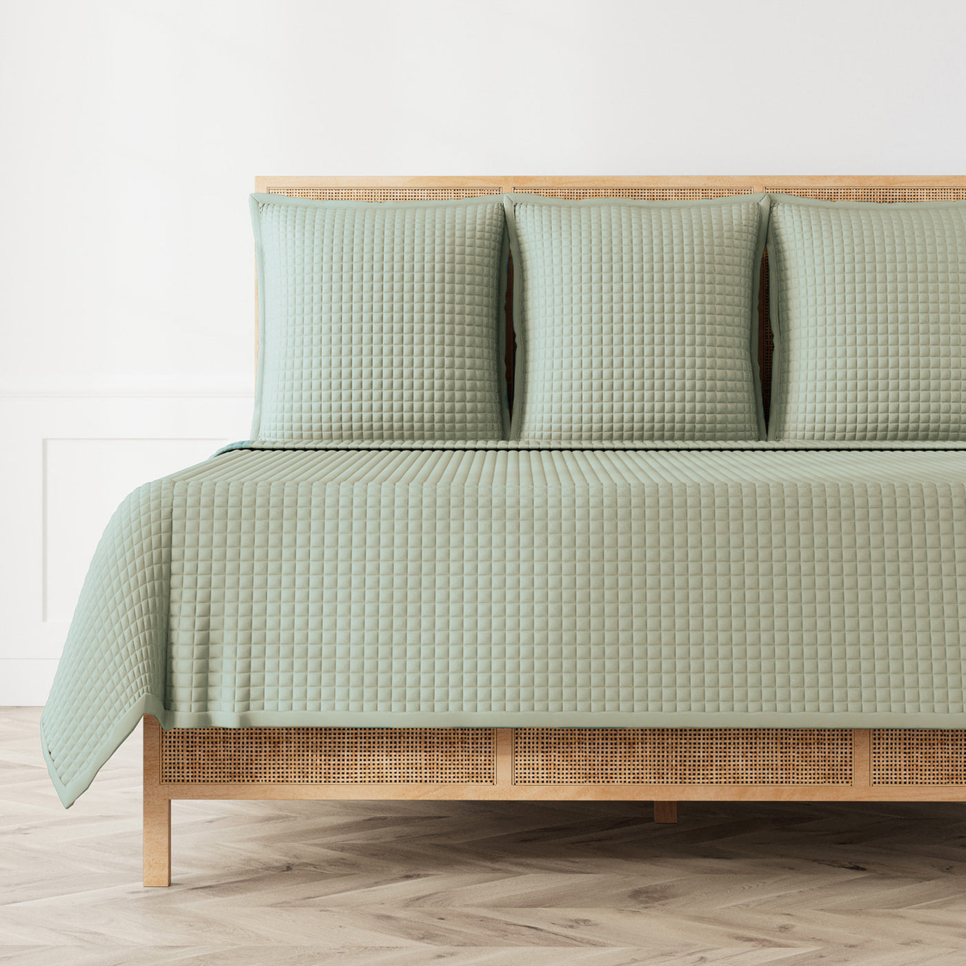 Sage green quilted coverlet and shams styled on a rattan bed frame, herringbone floor.