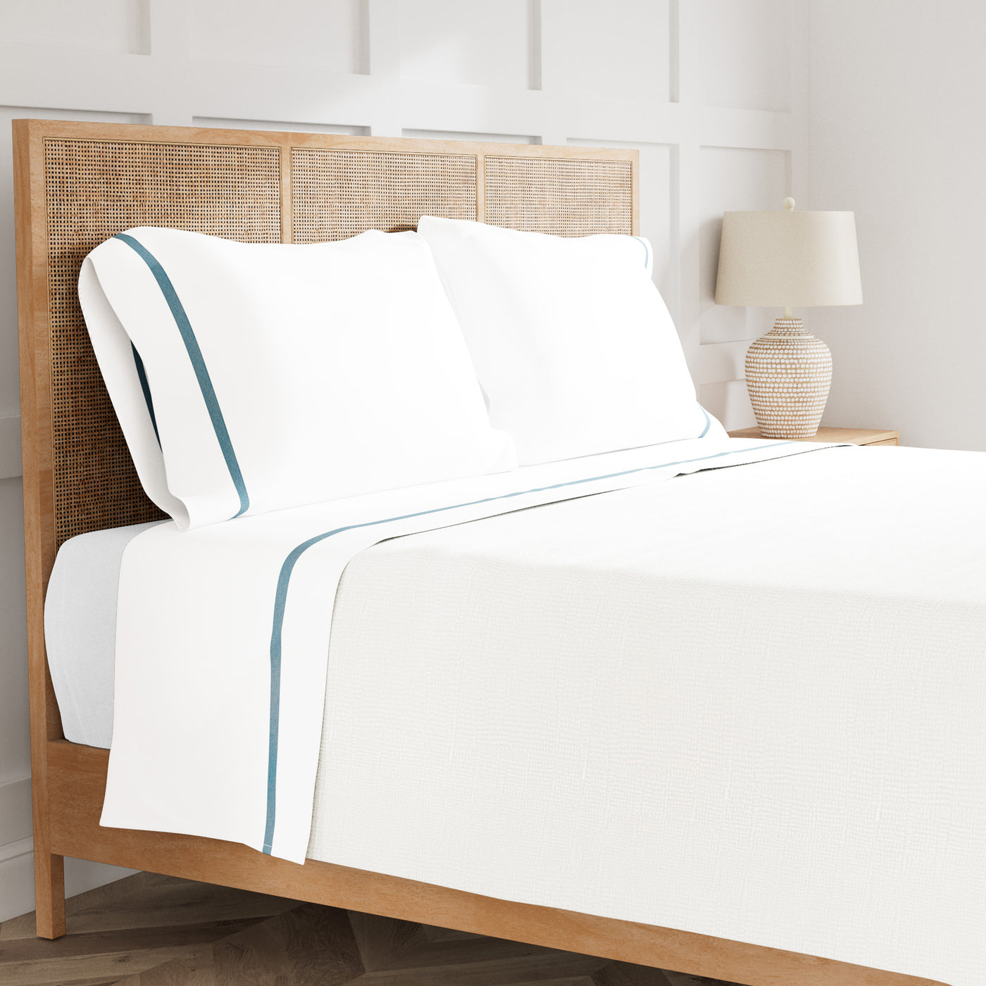 White cotton sheet set with teal satin trim, styled on a bed with rattan headboard.