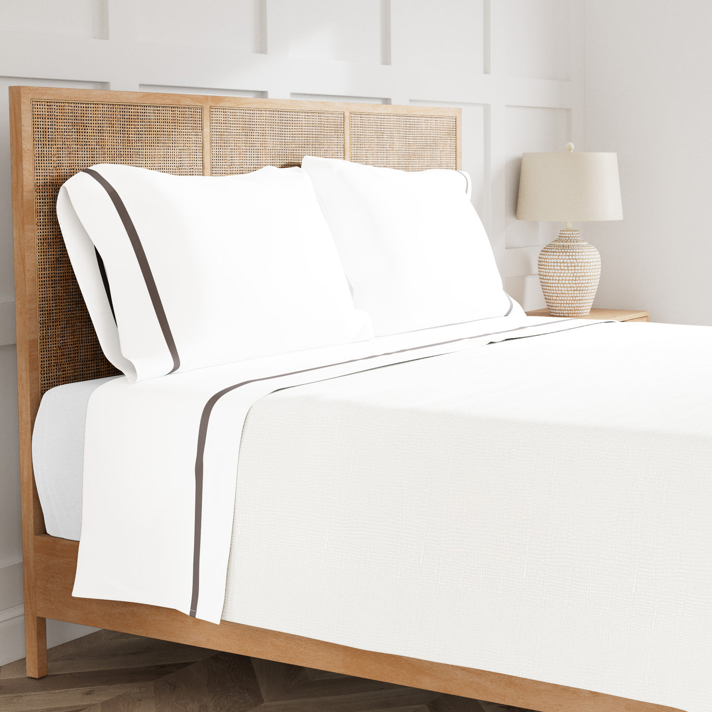 White cotton sheet set with taupe trim detail, styled on rattan bed with bedside lamp.