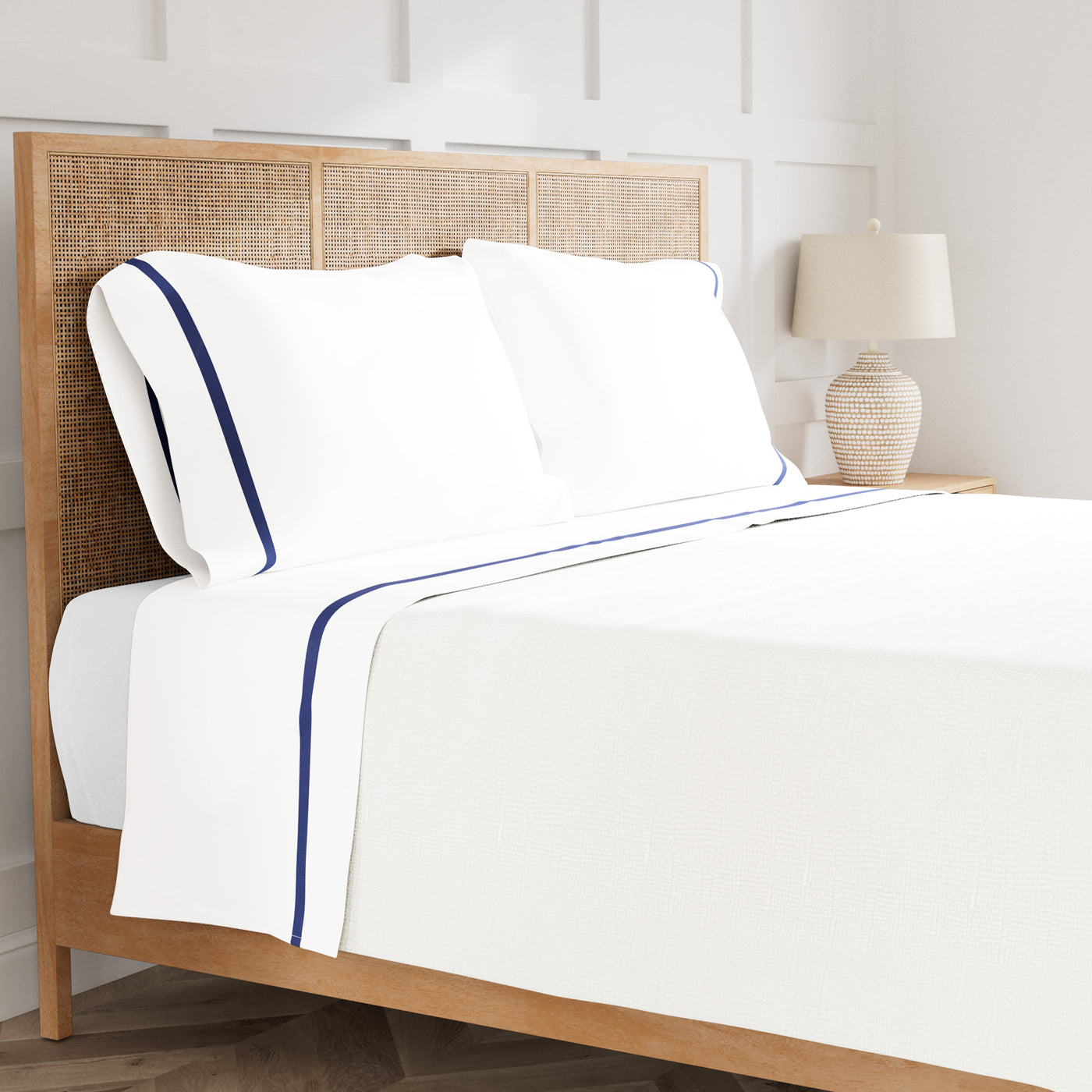 White sheet set with navy trim detail, styled on a rattan bed in a bright bedroom.