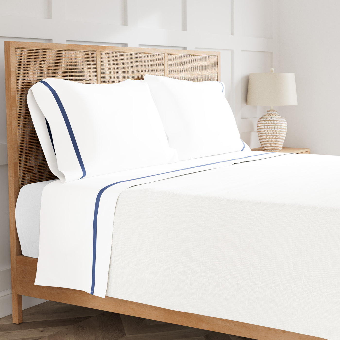 White cotton sheet set with navy trim, styled on a rattan bed in a bright bedroom setting.