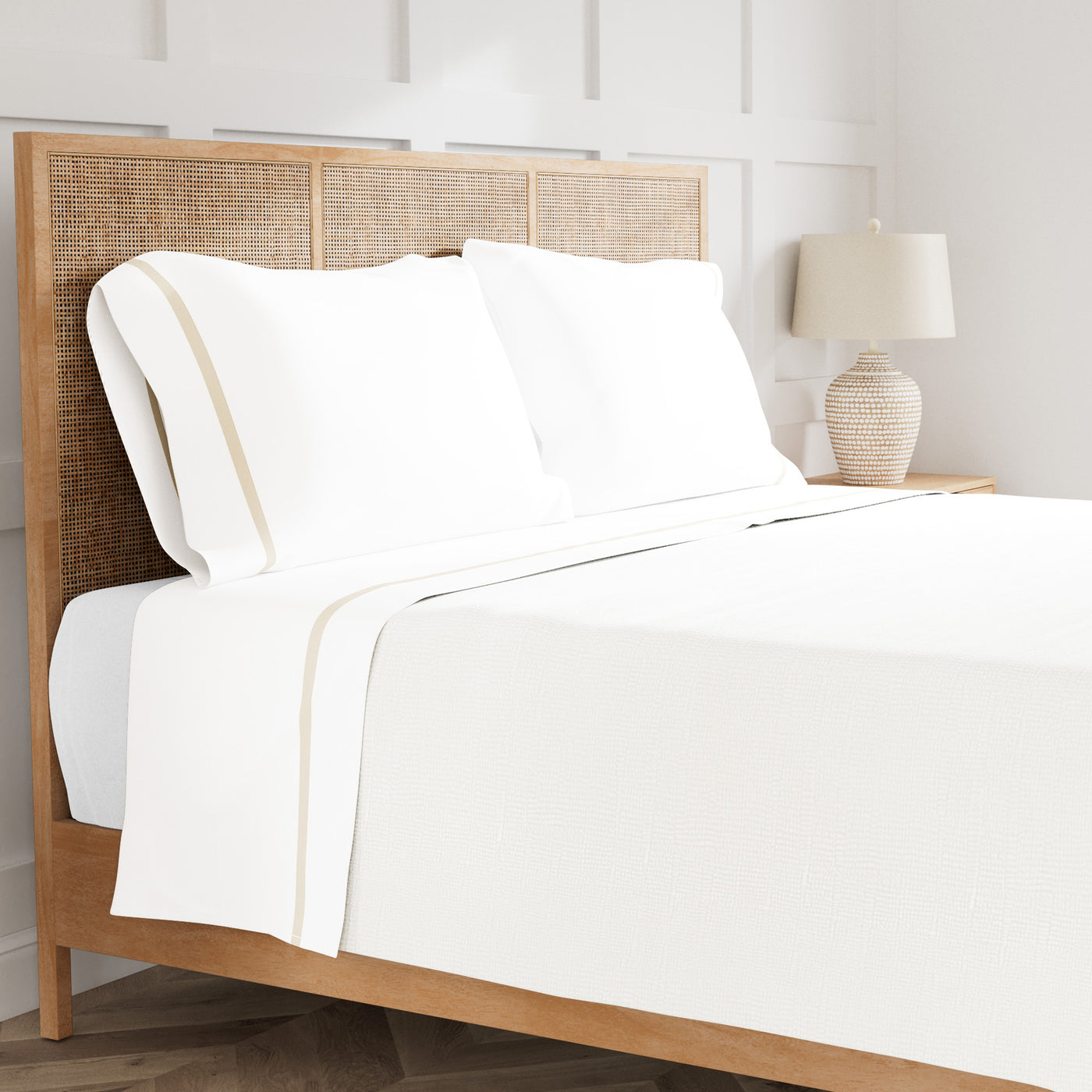White cotton sheet set with champagne trim, styled on a rattan bed in a bright bedroom.
