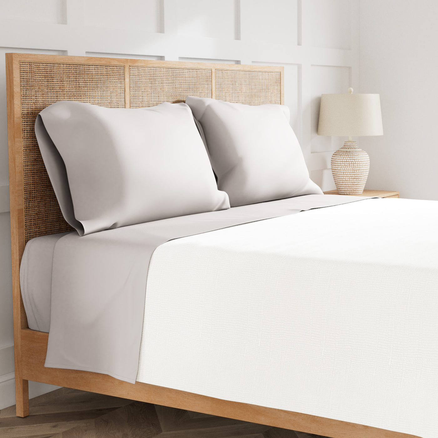 Pillowcases in silver-grey paired with white sheets on a rattan bed in a bright bedroom.