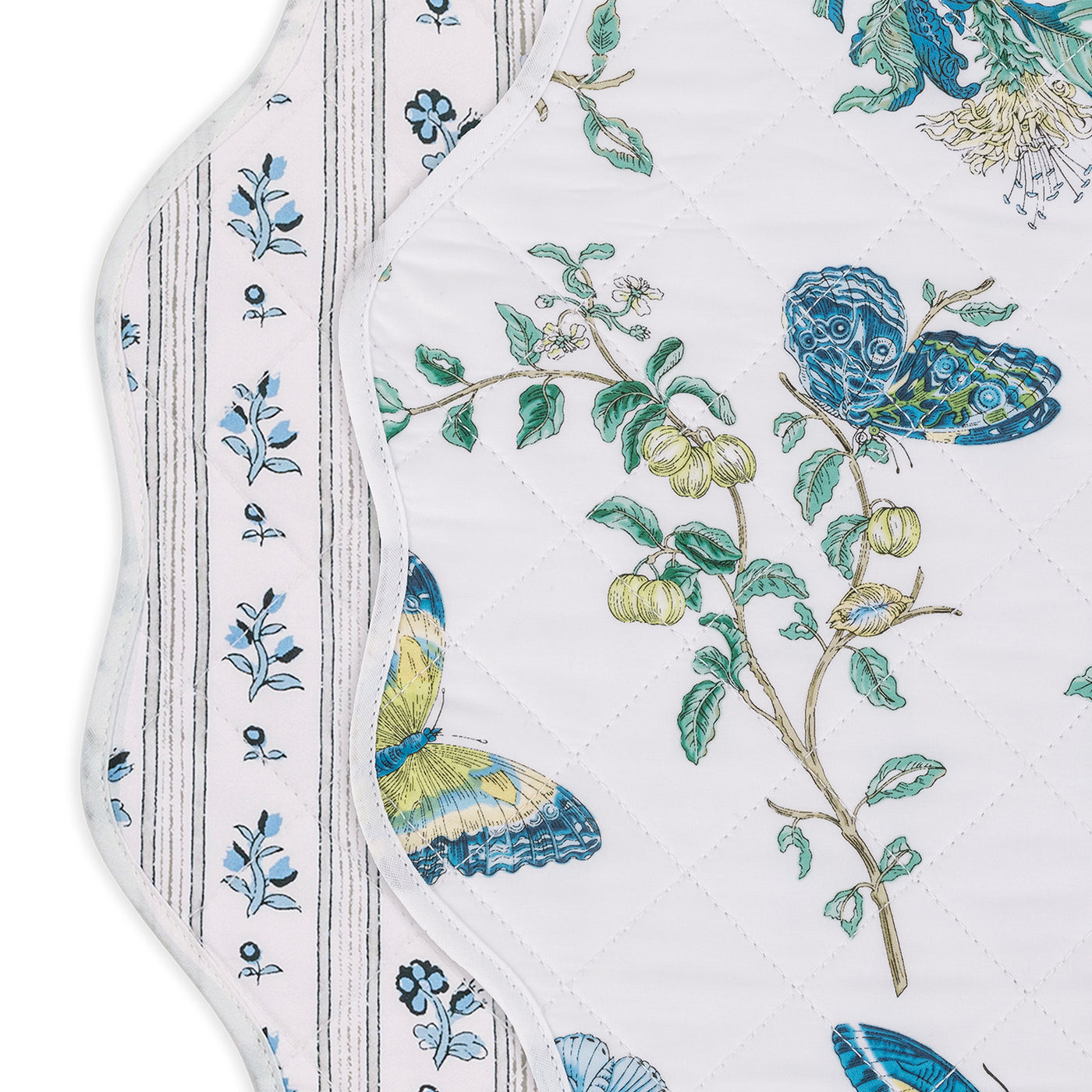 The Matouk Quilted Reversible Placemat Set includes two scalloped placemats—one with blue floral stripes, the other with blue and yellow butterflies, green branches, and white blossoms—ideal for stylish dining table decor.