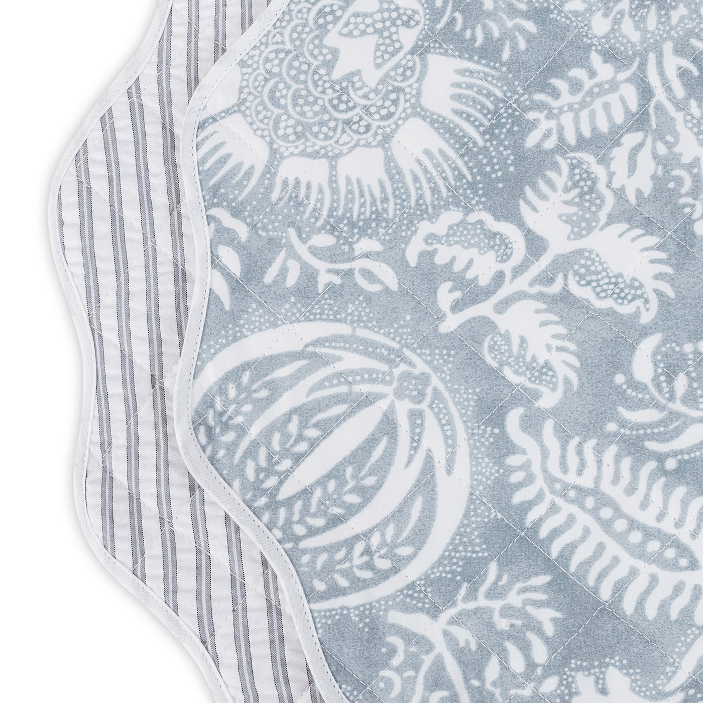 Close-up of two Matouk Quilted Reversible Placemats—one with gray and white florals, the other with stripes. Both have scalloped edges, making this set ideal for stylish table decor.
