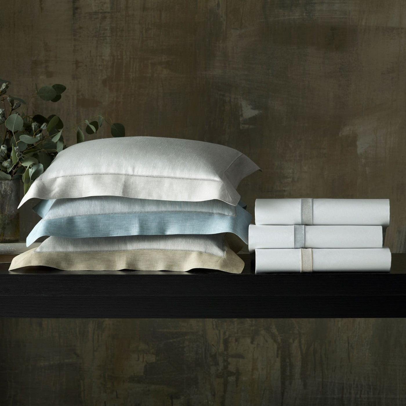 Stacked pillowcases in ivory, sky blue, and champagne beside folded white sheets on a dark shelf.