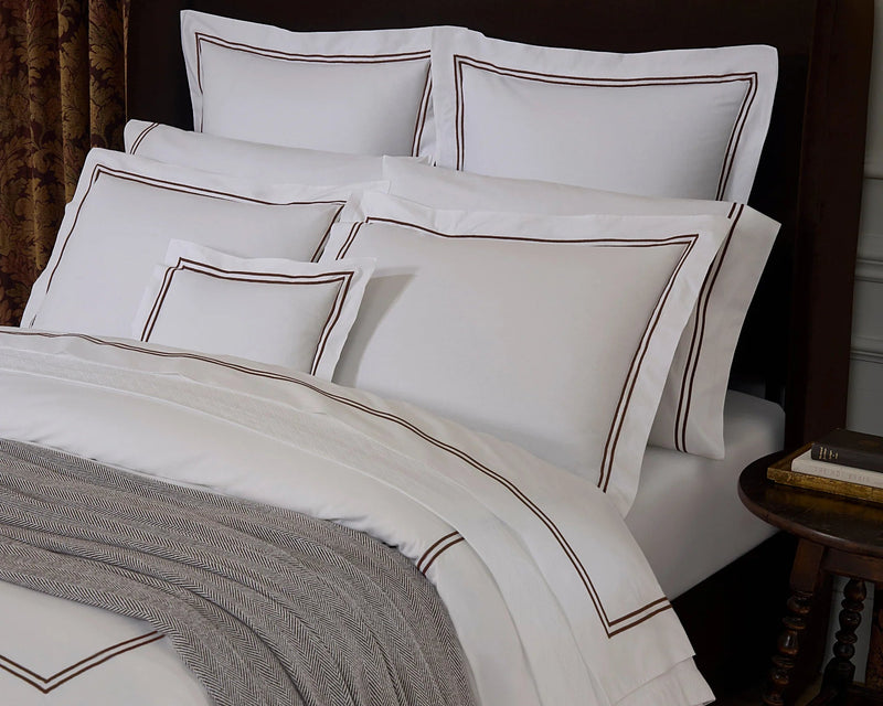 White bedding set with brown embroidered border details, styled on a bed with a dark brown headboard, a gray throw blanket, and a bedside table in a bedroom setting.