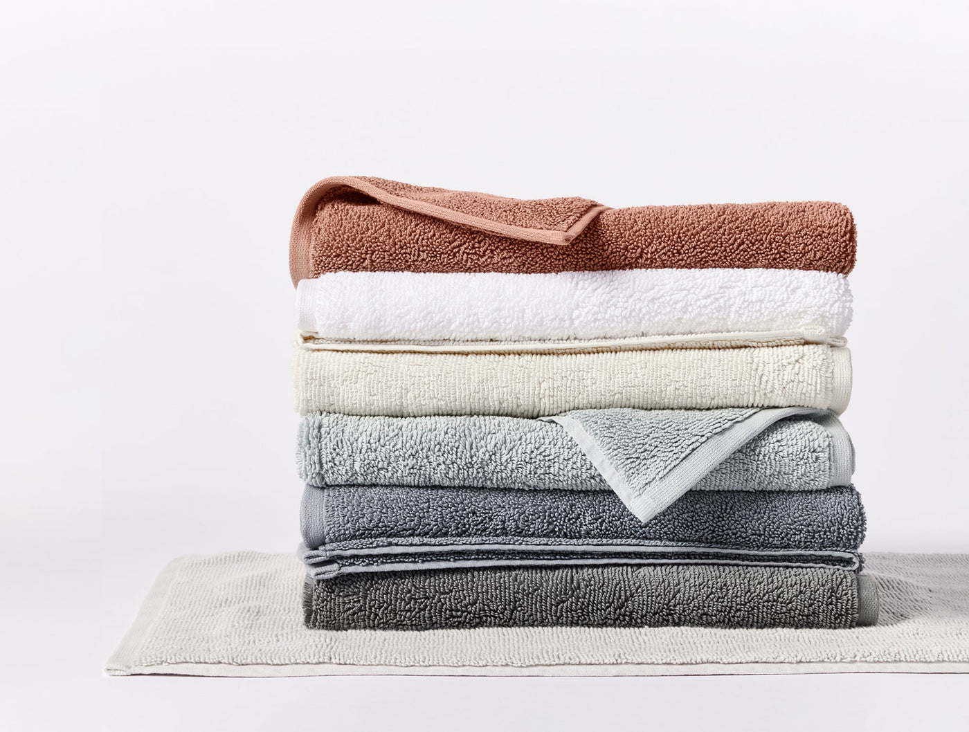 Stack of folded Coyuchi Cloud Loom towels in terracotta, white, sage, and slate on a bath rug.
