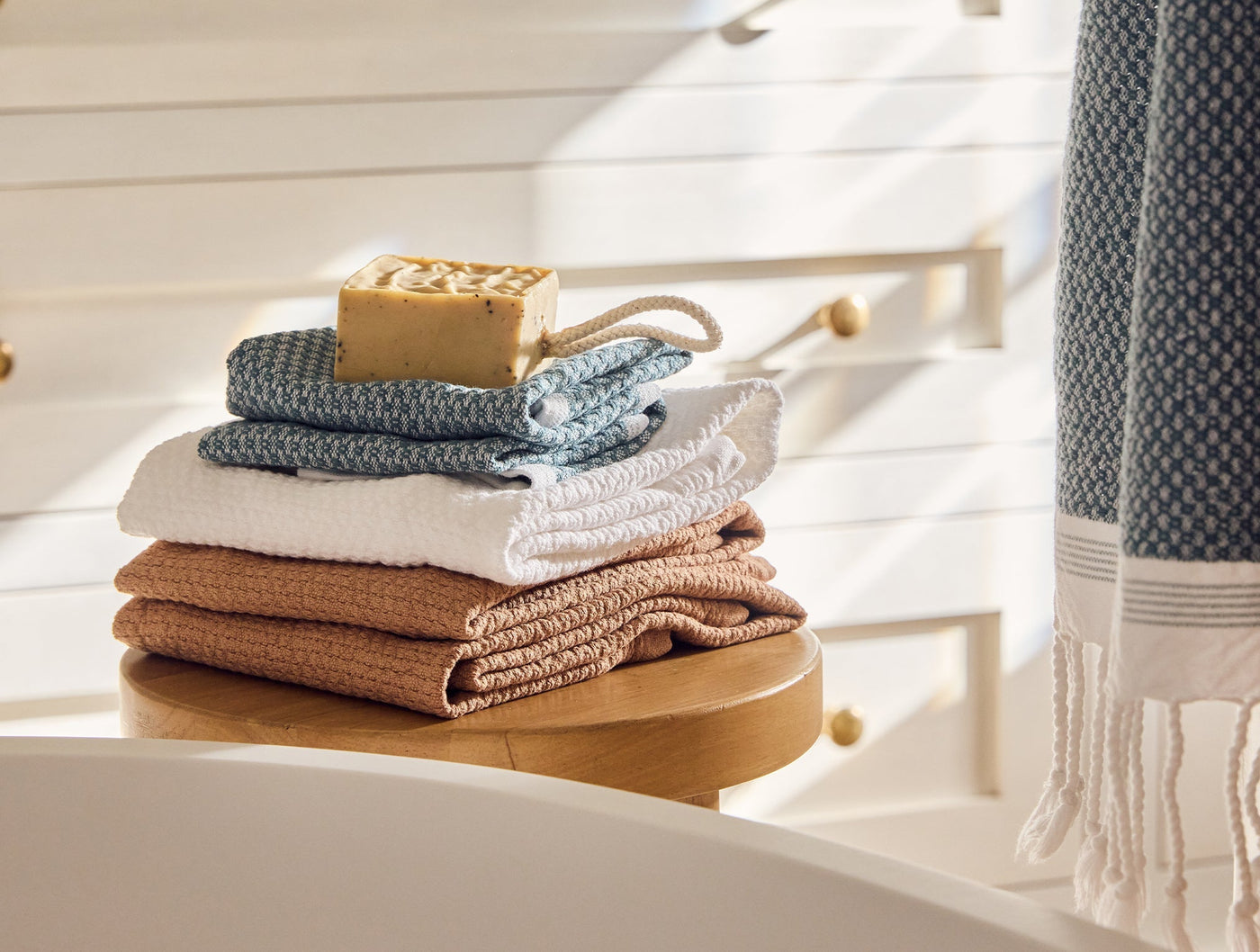 Stack of folded Coyuchi Adriatic towels in slate, white, and terracotta with waffle texture, styled in sunlit bathroom.