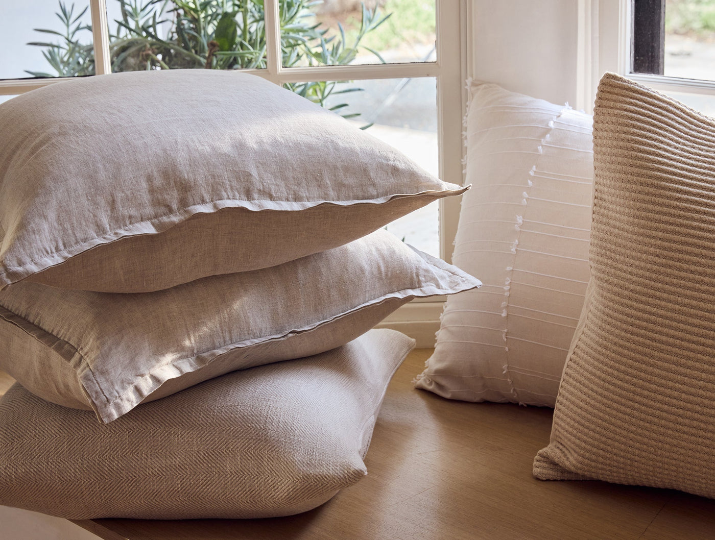 Close-up of stacked natural linen Coyuchi pillow covers beside a ribbed camel cushion near a window.