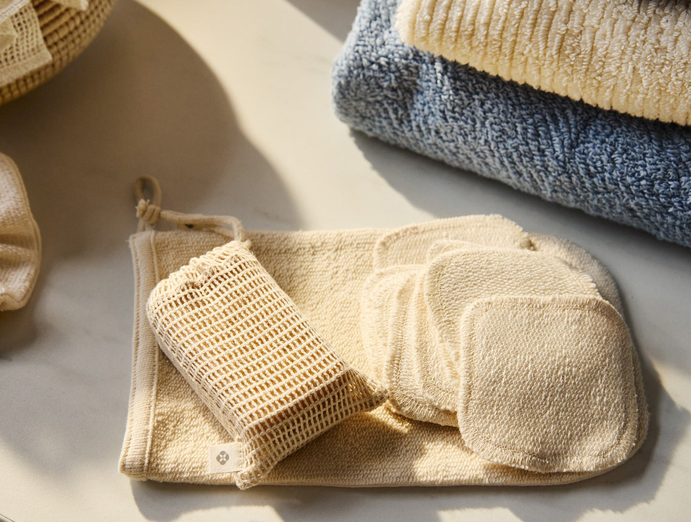 Coyuchi Bare soap bag with face pads and wash cloth, blue towels stacked in background.