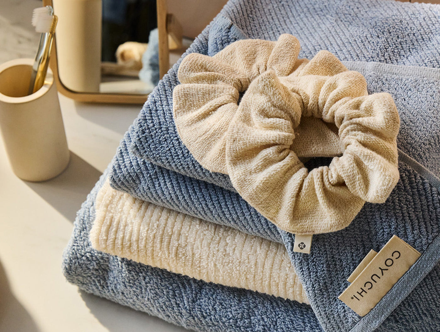 Coyuchi Bare scrunchie set in sand tones resting on folded blue textured towels.