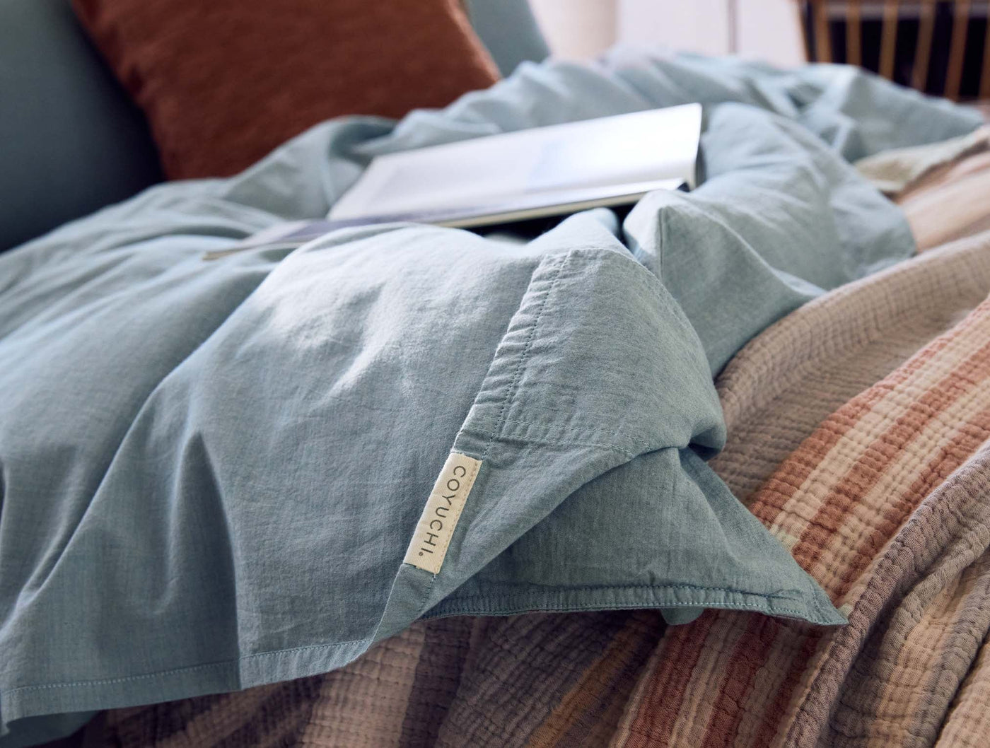 Coyuchi crinkled percale pillowcase in slate blue, casually draped on styled bed with open book.