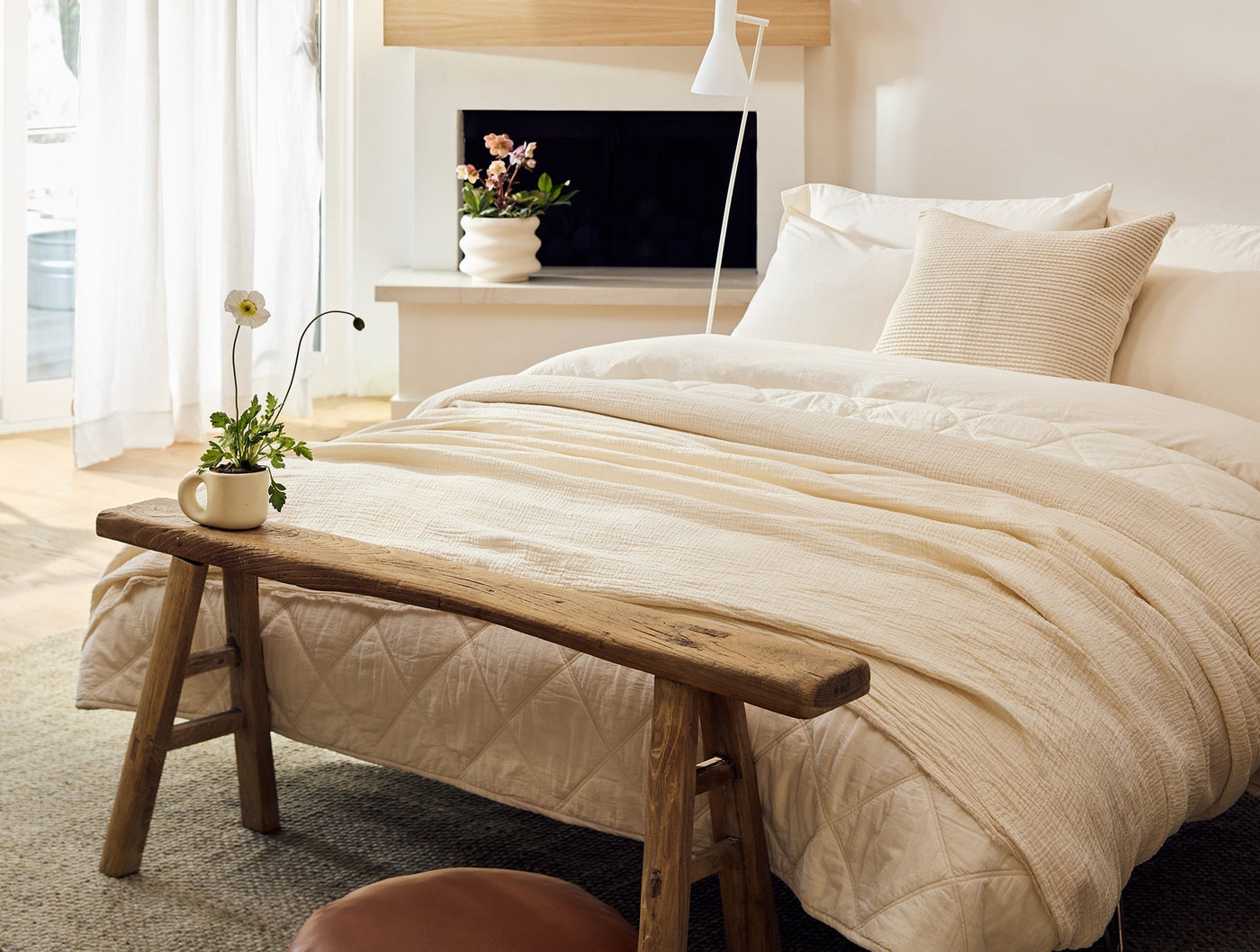 Ivory matelassé blanket draped over bed, rustic wood bench in foreground, bright airy room.
