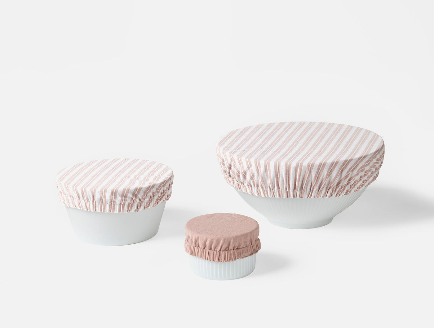 Three Coyuchi bowl covers in blush stripe and solid blush, covering white bowls in small, medium, large sizes.