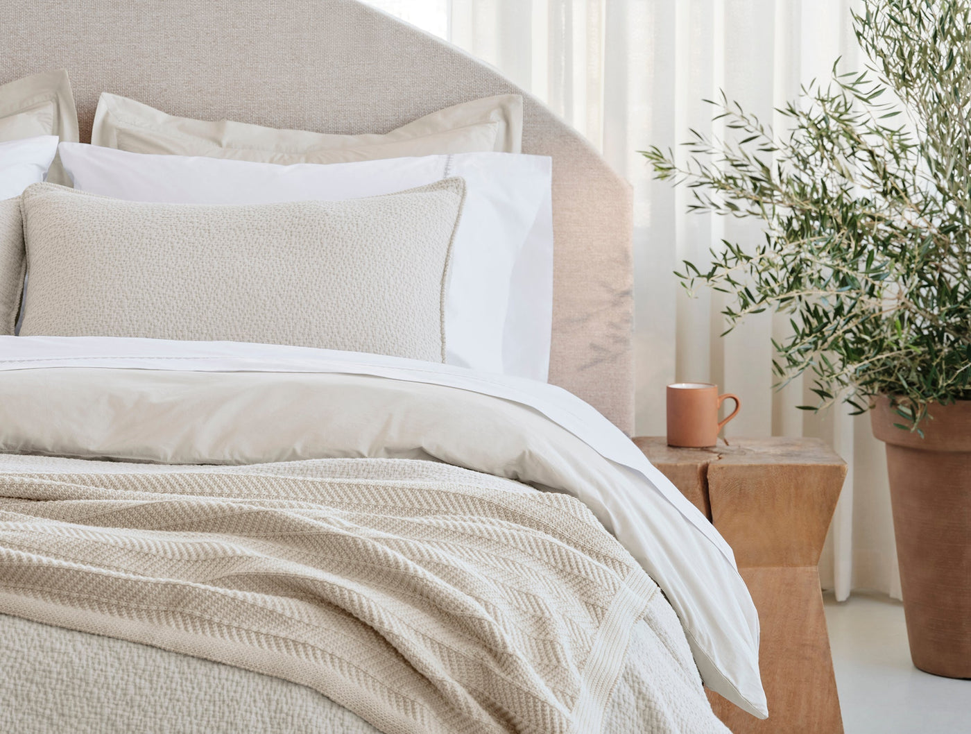 Ivory embroidered pillow sham layered with white sheets on a styled bed, olive tree beside.