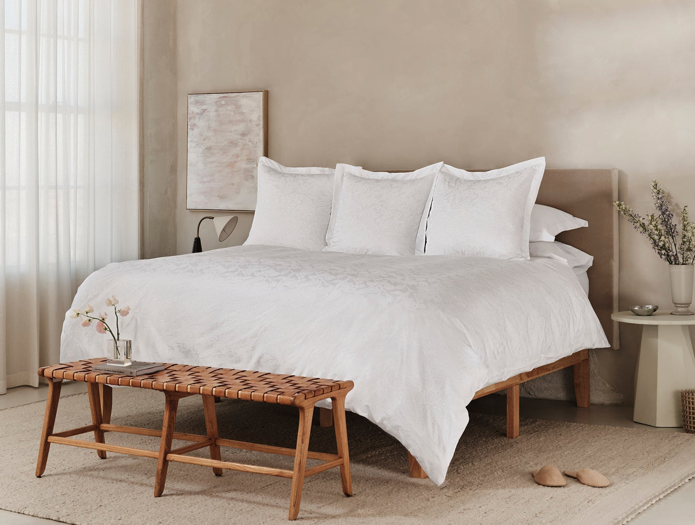 White jacquard bedding styled on a bed with woven bench and neutral bedroom setting.