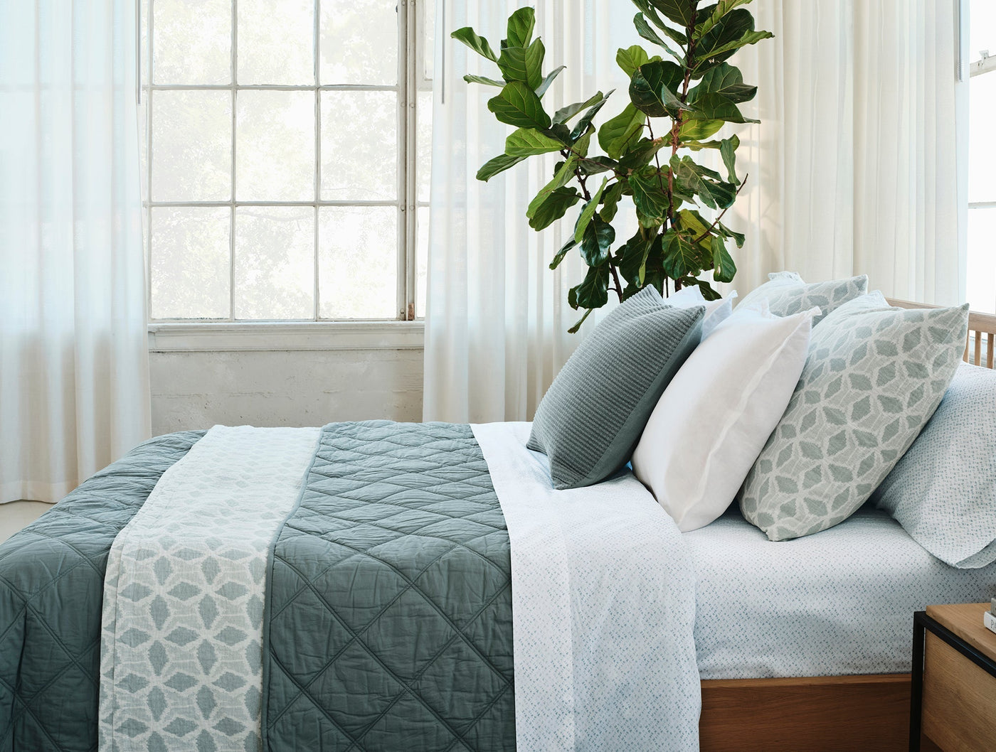 Quilted sage green blanket draped over a bed with layered pillows near a sunlit window.
