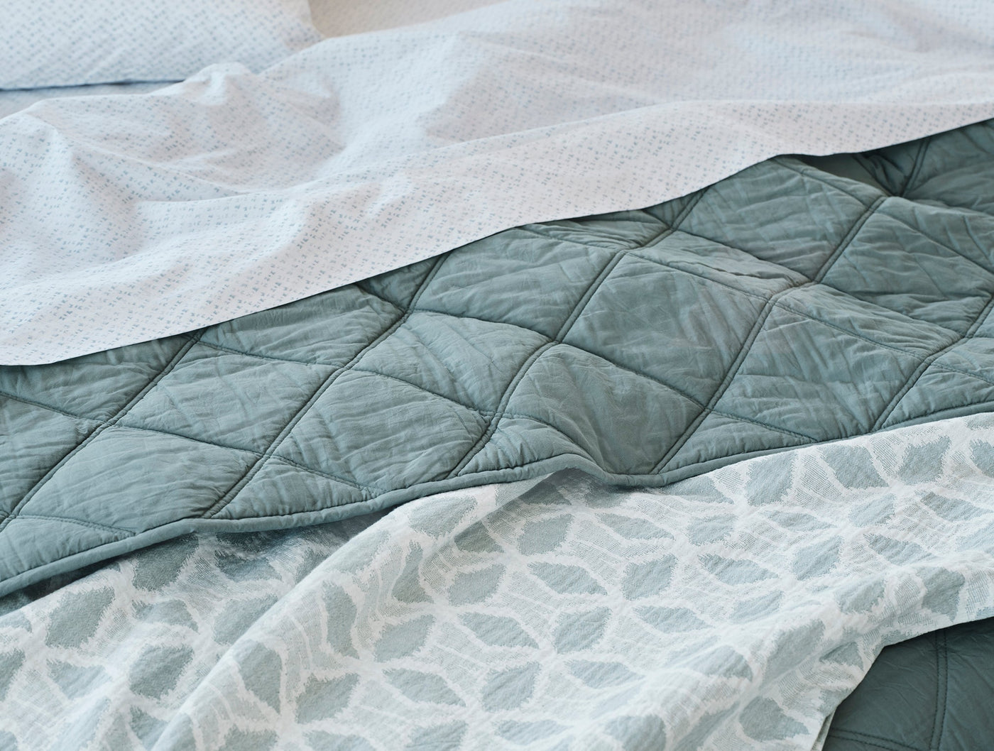 Close-up of teal quilted diamond-pattern blanket layered with leaf-print and white textured bedding.