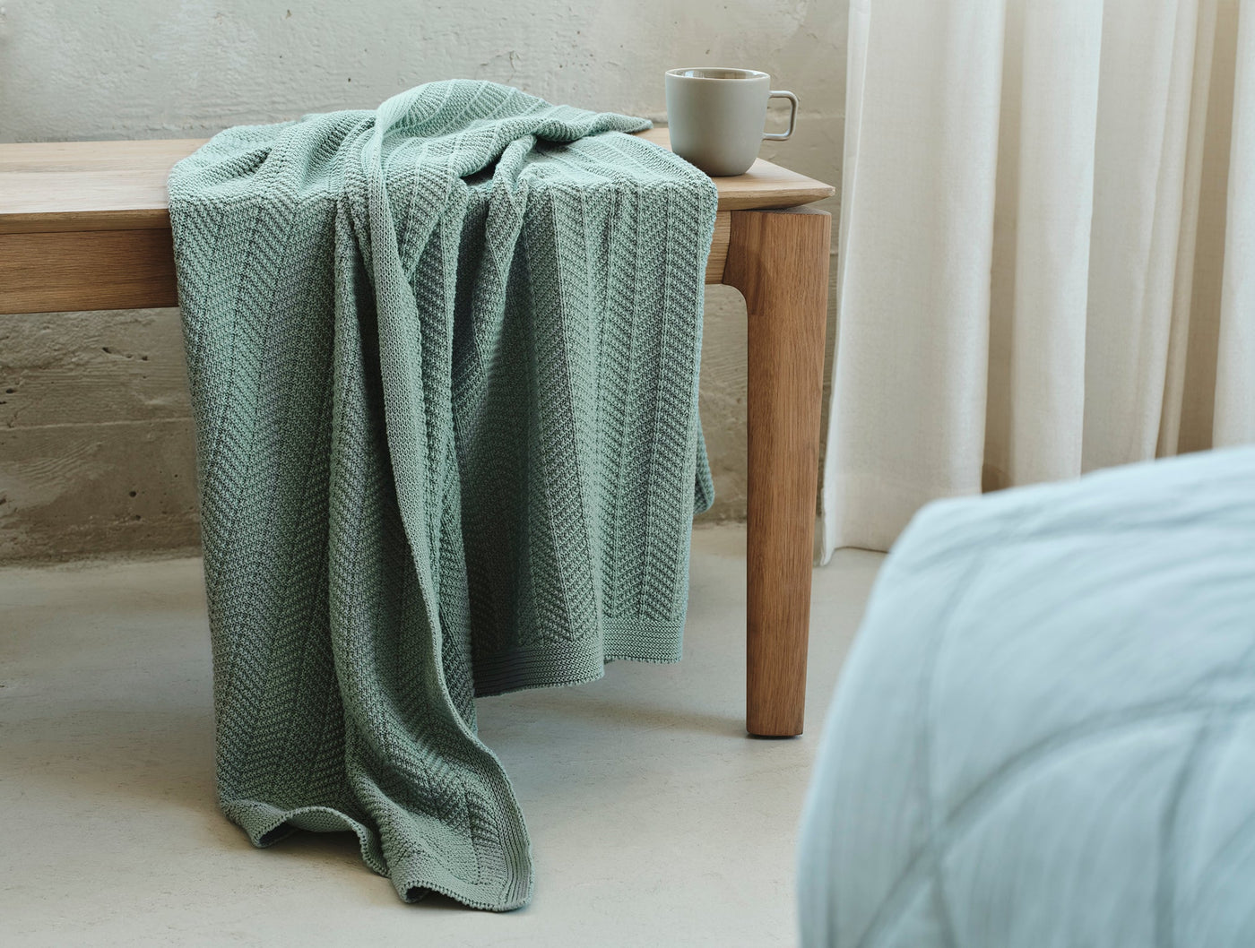 Coyuchi Hermosa Knit Throw in sage green draped over a wooden bench with a mug.