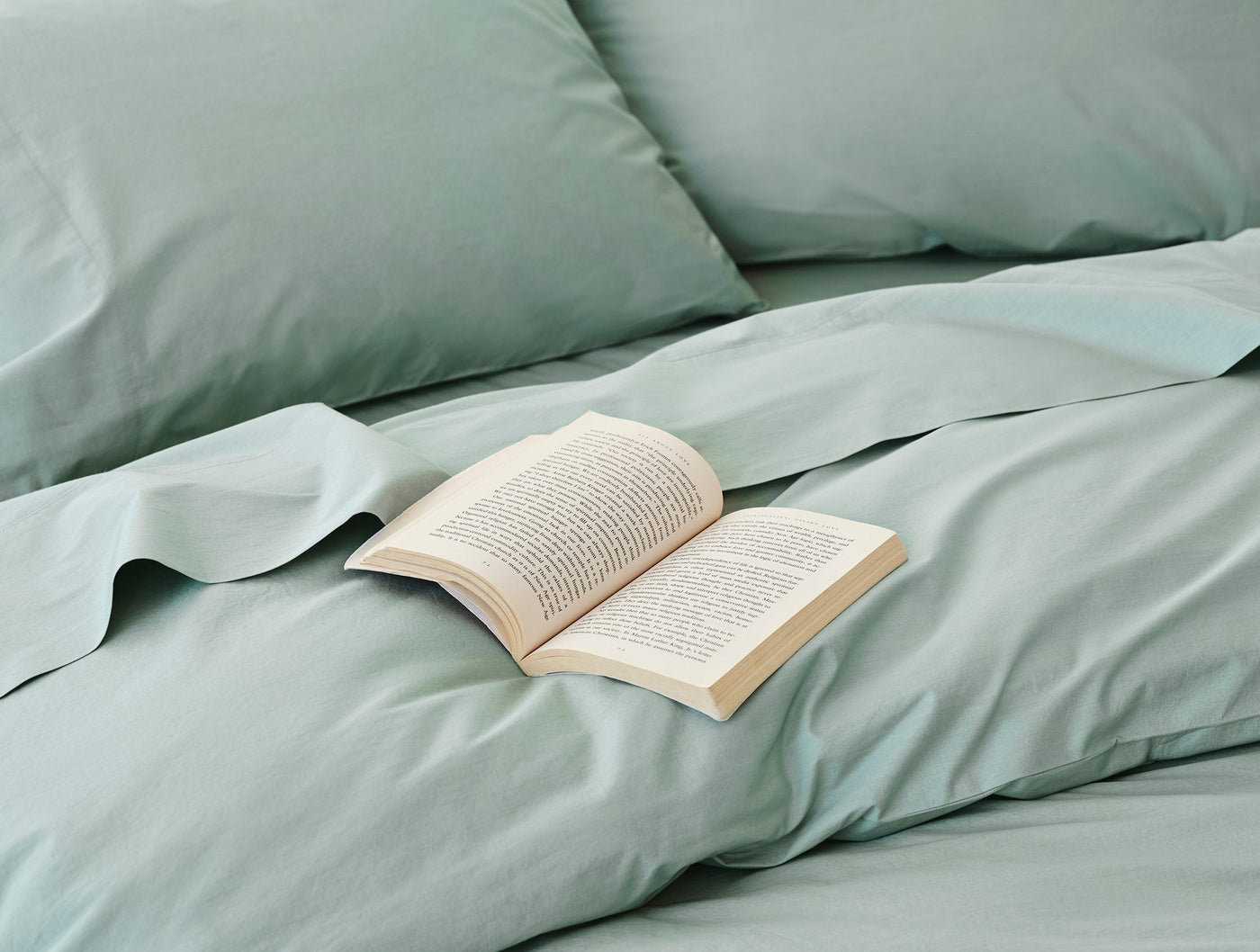 Sage green percale sheet set styled on bed with open book resting on duvet.