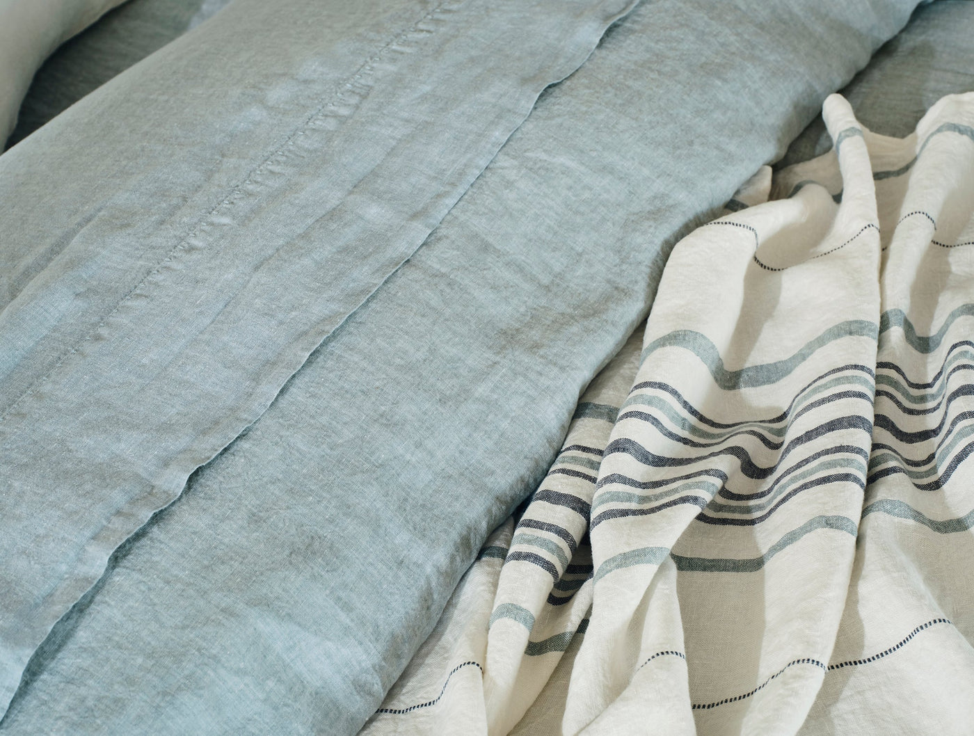 Close-up of sage linen sheet alongside ivory linen with slate stripe detail, draped and folded.