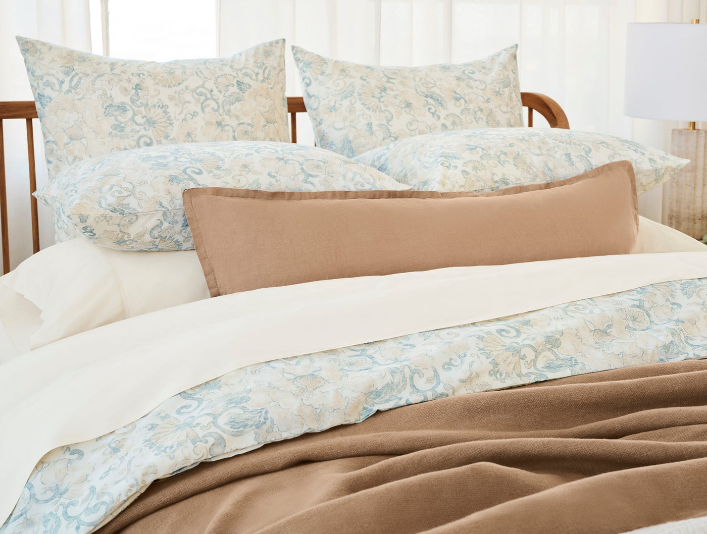 Ivory and blue floral print shams styled on a bed with camel bolster and blanket.
