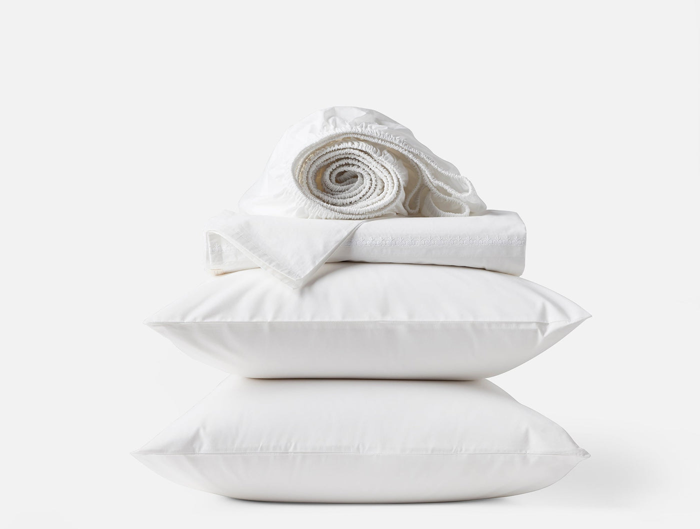 Coyuchi Floret Embroidered sheet set in white, stacked with pillows and rolled sheets.