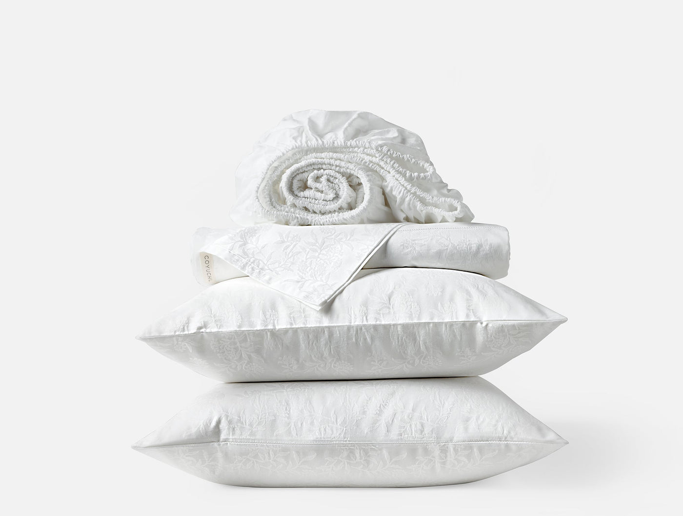 Coyuchi Heirloom Sateen Jacquard sheet set in white, stacked with pillows and rolled fitted sheet.