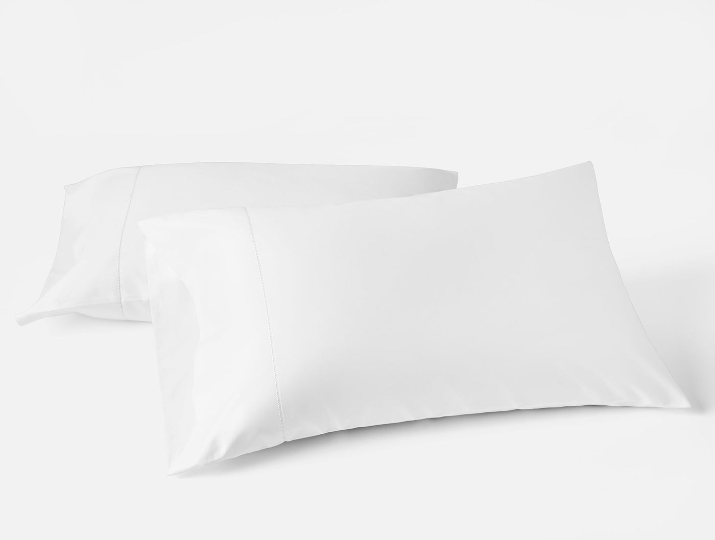Close-up of two white Coyuchi Heirloom Sateen pillowcases arranged overlapping on white surface.