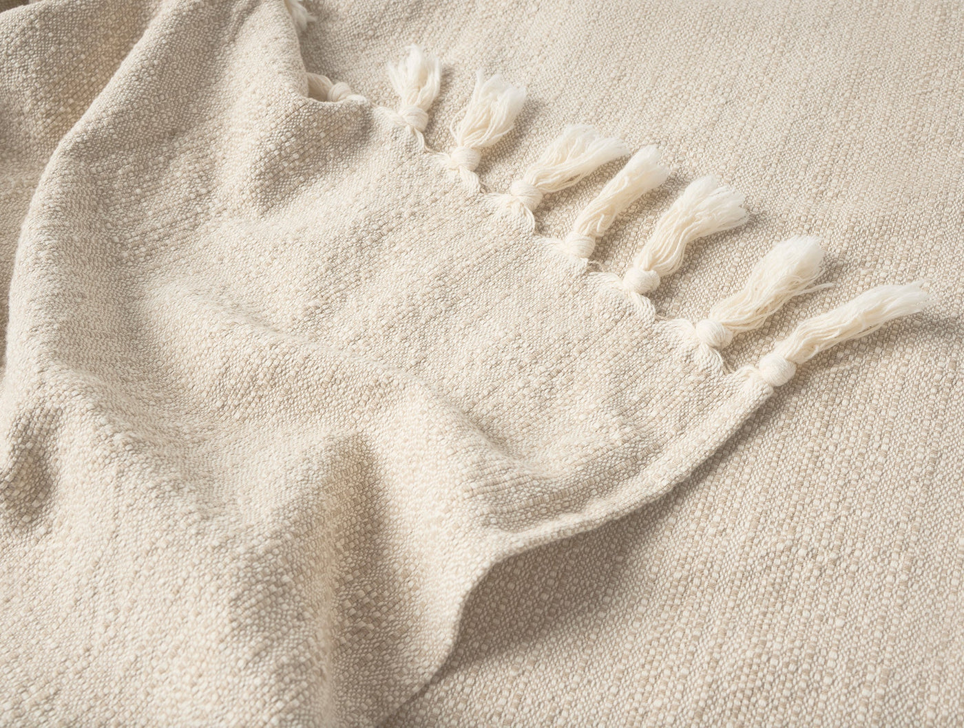 Close-up of ivory woven throw corner with knotted fringe detail against textured surface.