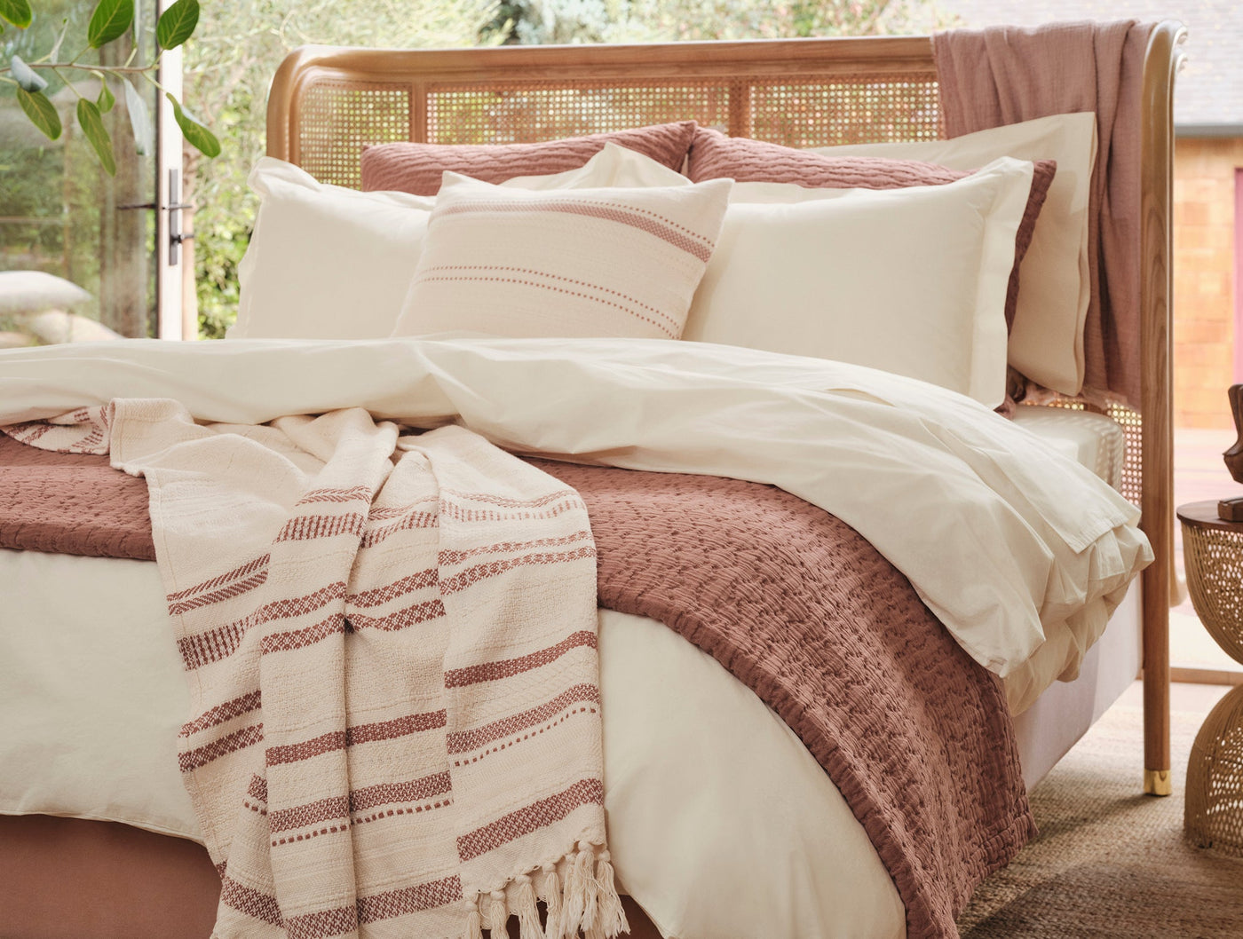 Ivory percale sheet set styled on rattan bed with dusty rose accents and layered bedding.