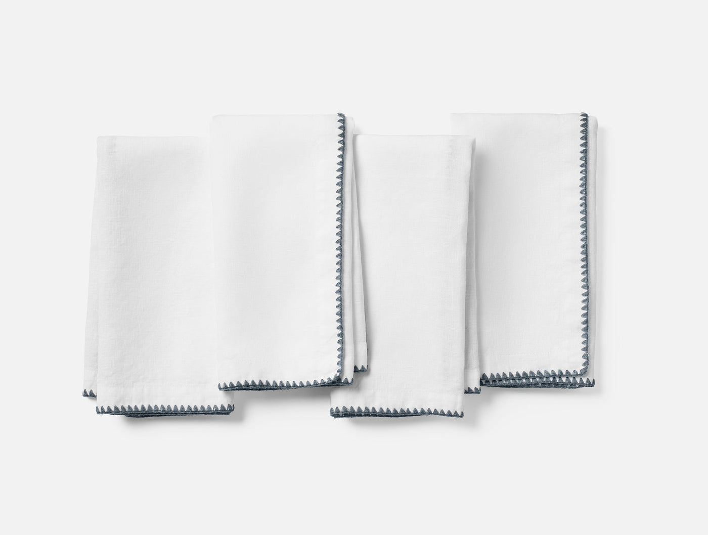 Close-up of four white linen napkins with slate blue whipstitch embroidered edges, folded flat.