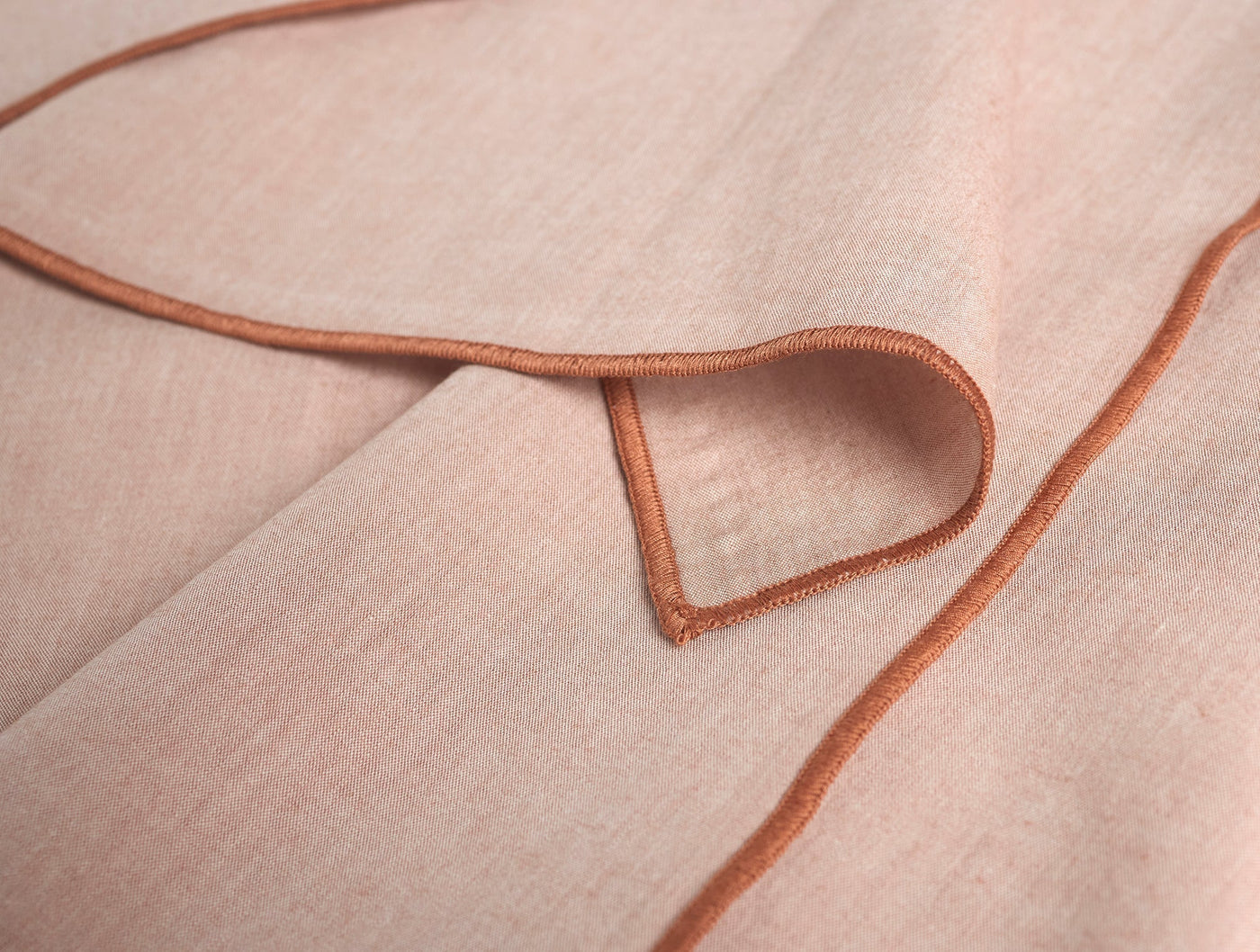 Close-up of blush percale napkin corner with terracotta satin-stitch border detail.