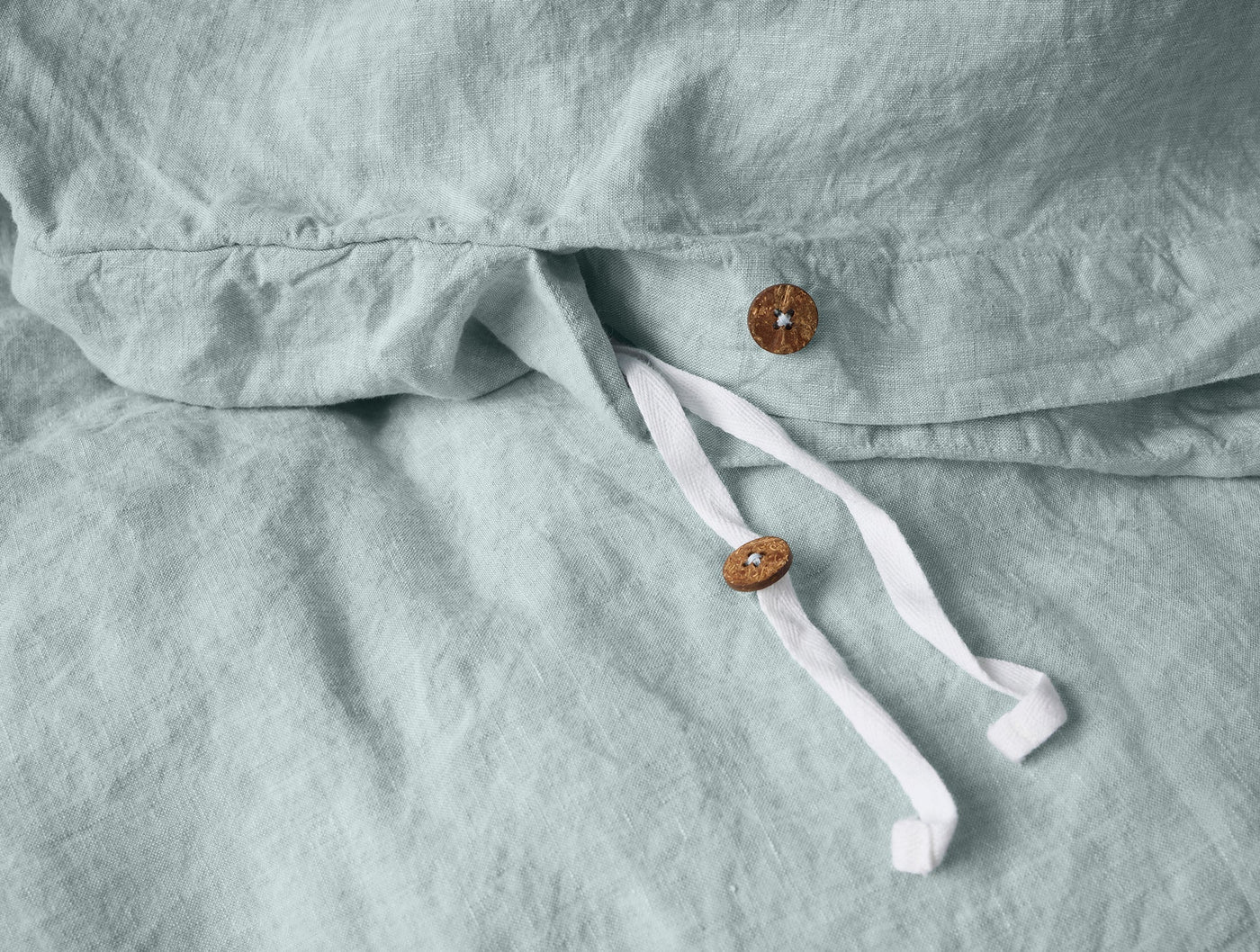 Close-up of sage linen duvet cover closure with two wooden buttons and white ribbon ties.