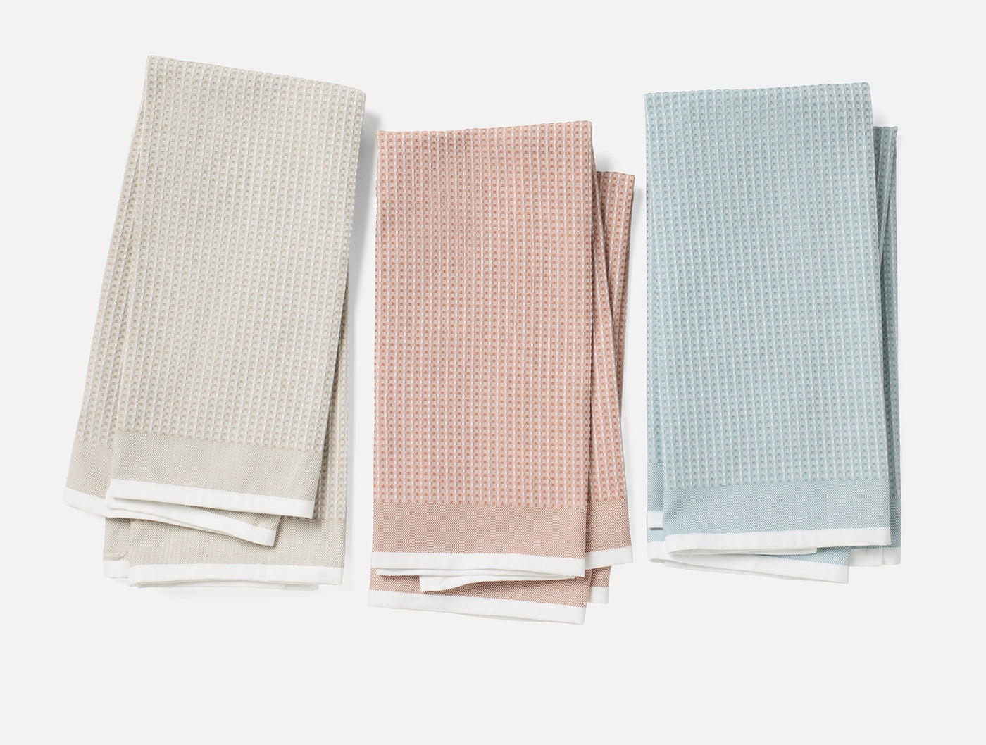 Three folded Coyuchi waffle-weave kitchen towels in stone, blush, and sky blue with white borders.