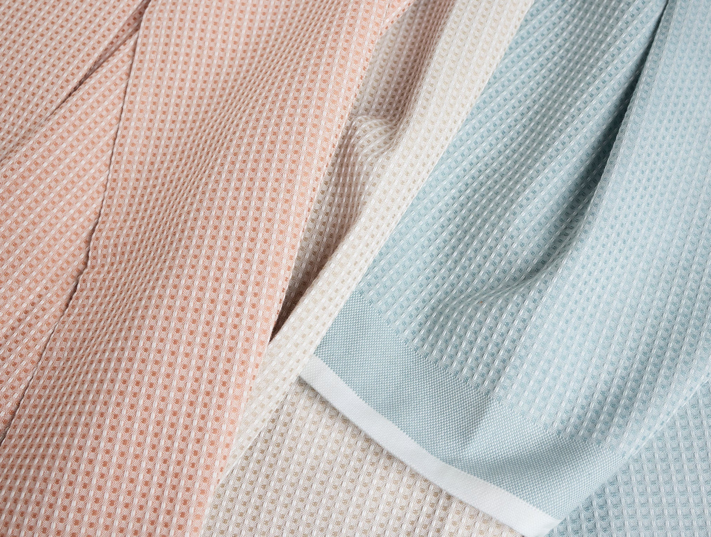 Close-up of Coyuchi waffle-knit kitchen towels in blush, ivory, and sky blue with white hem.