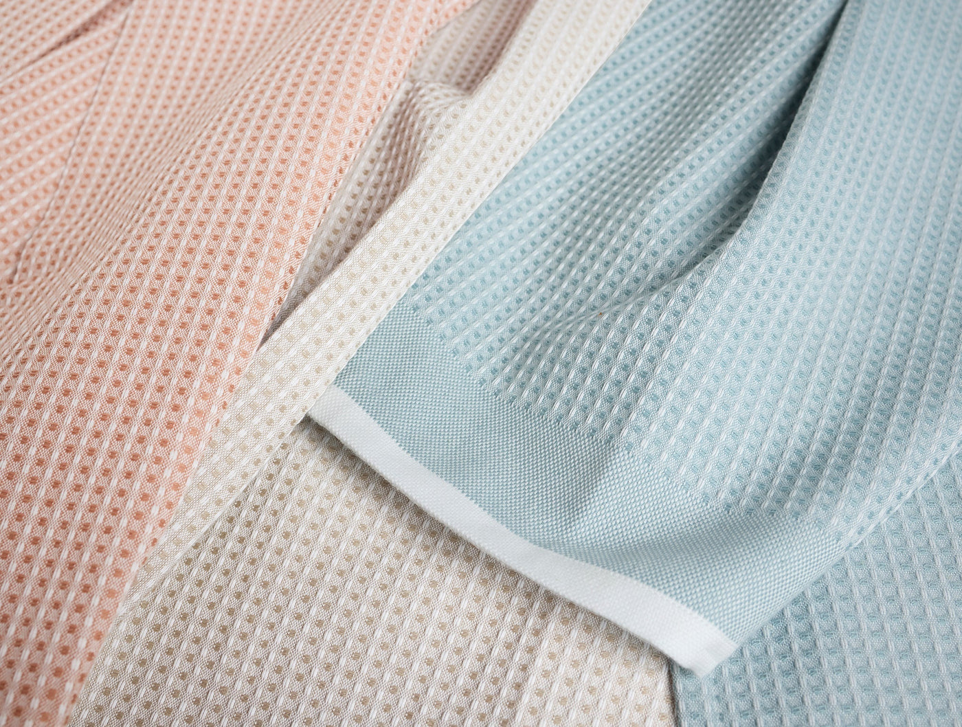 Close-up of Coyuchi waffle-knit kitchen towels in blush, ivory, and mist with white hem trim.