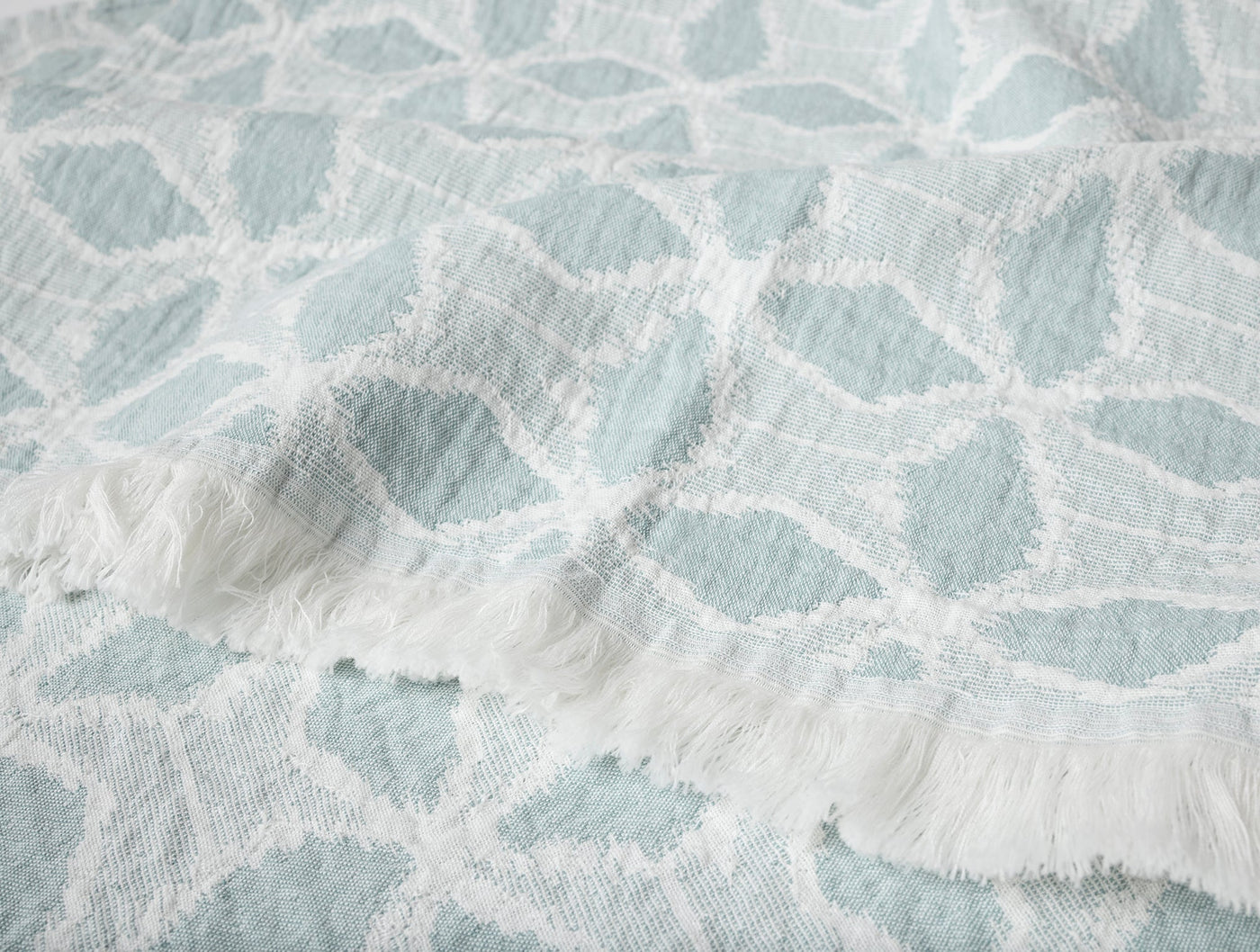 Close-up of Coyuchi Palomar throw fringe edge, seafoam jacquard leaf pattern on white.