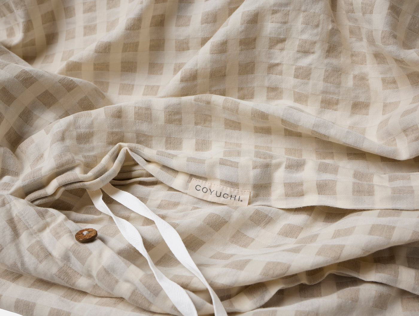 Close-up of Coyuchi Birch duvet cover corner showing label, tie, and button in oat gingham.