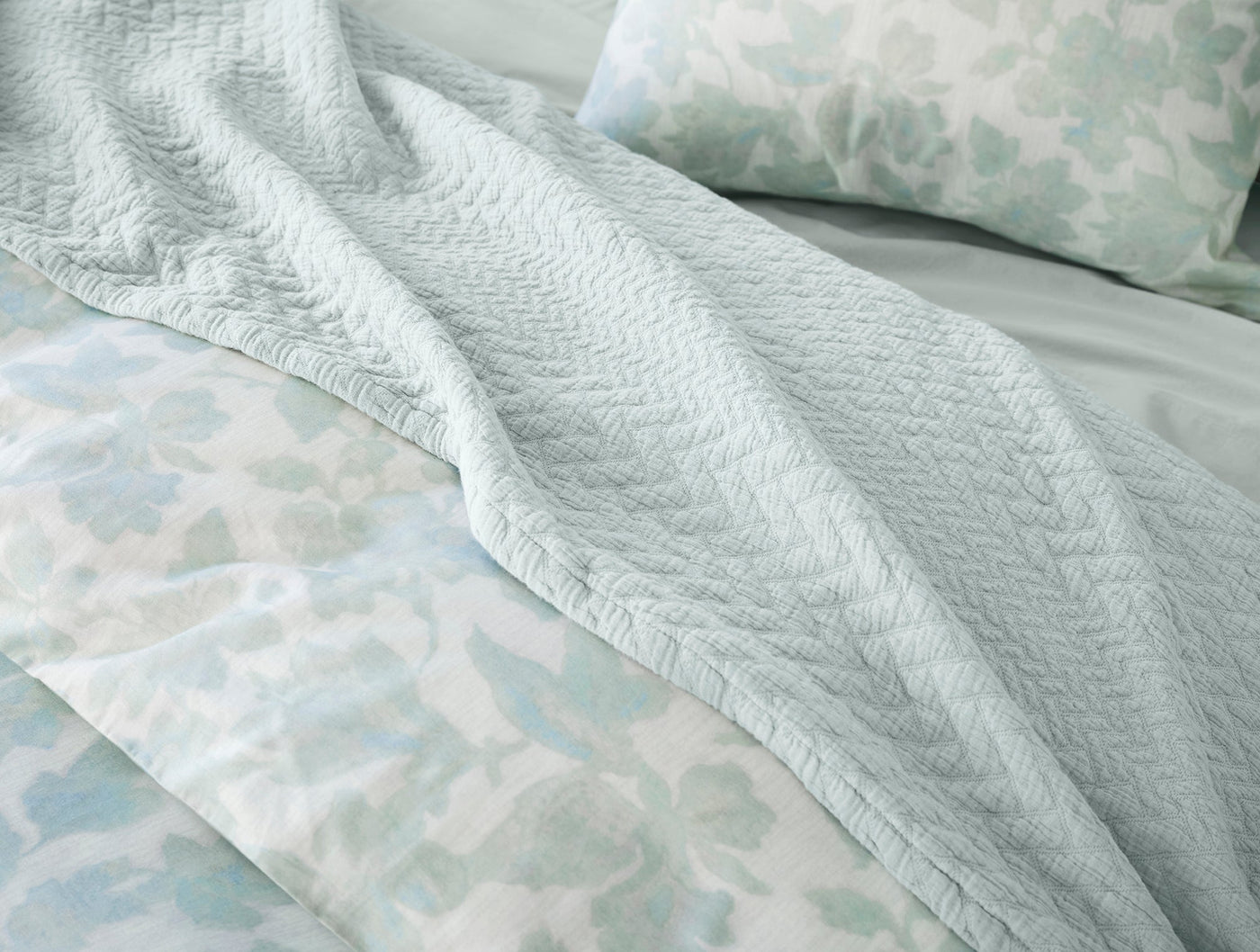 Close-up of Coyuchi Angeles Matelassé blanket in seafoam with herringbone texture draped over floral bedding.