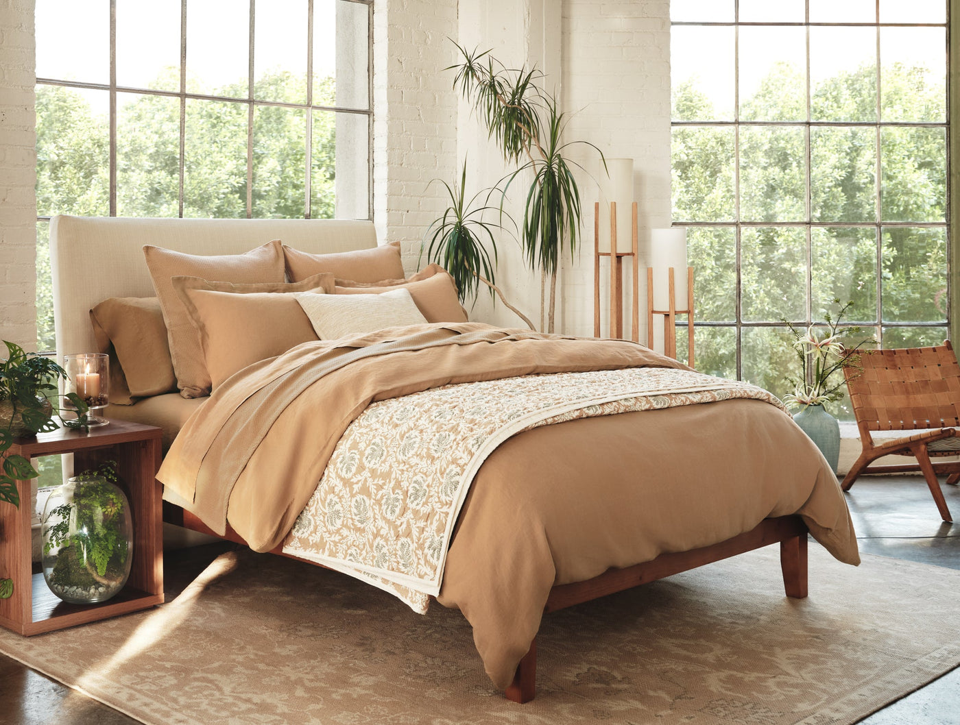 Camel linen sheet set styled on bed with floral runner, natural light, plants in background.