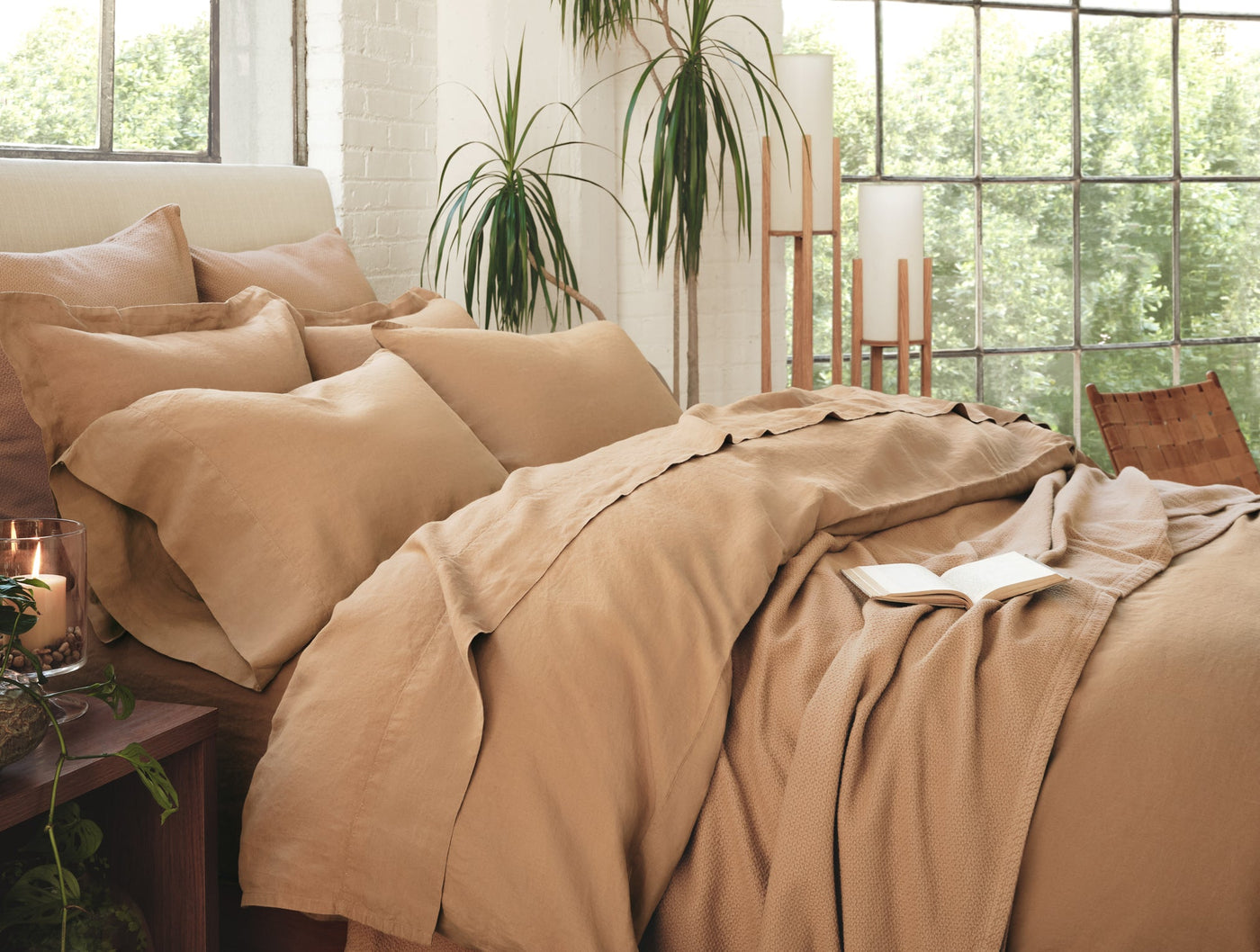 Caramel Coyuchi Relaxed Linen sheet set draped on a bed in a sun-lit room with plants.