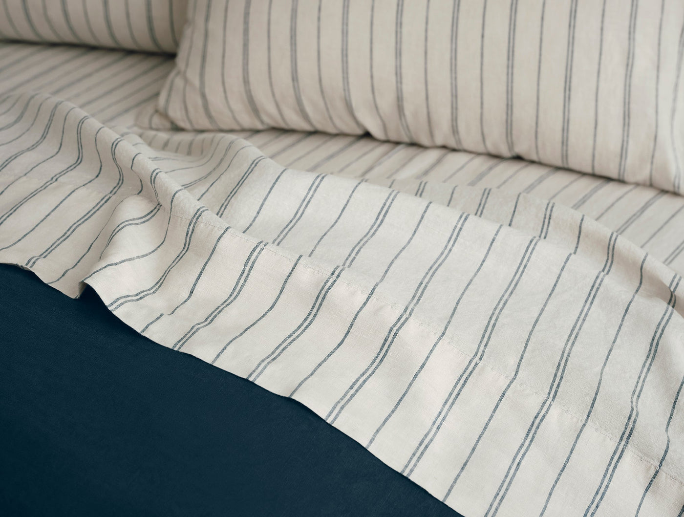 Close-up of ivory Coyuchi Relaxed Linen sheet with slate blue stripes draped over navy bedding.