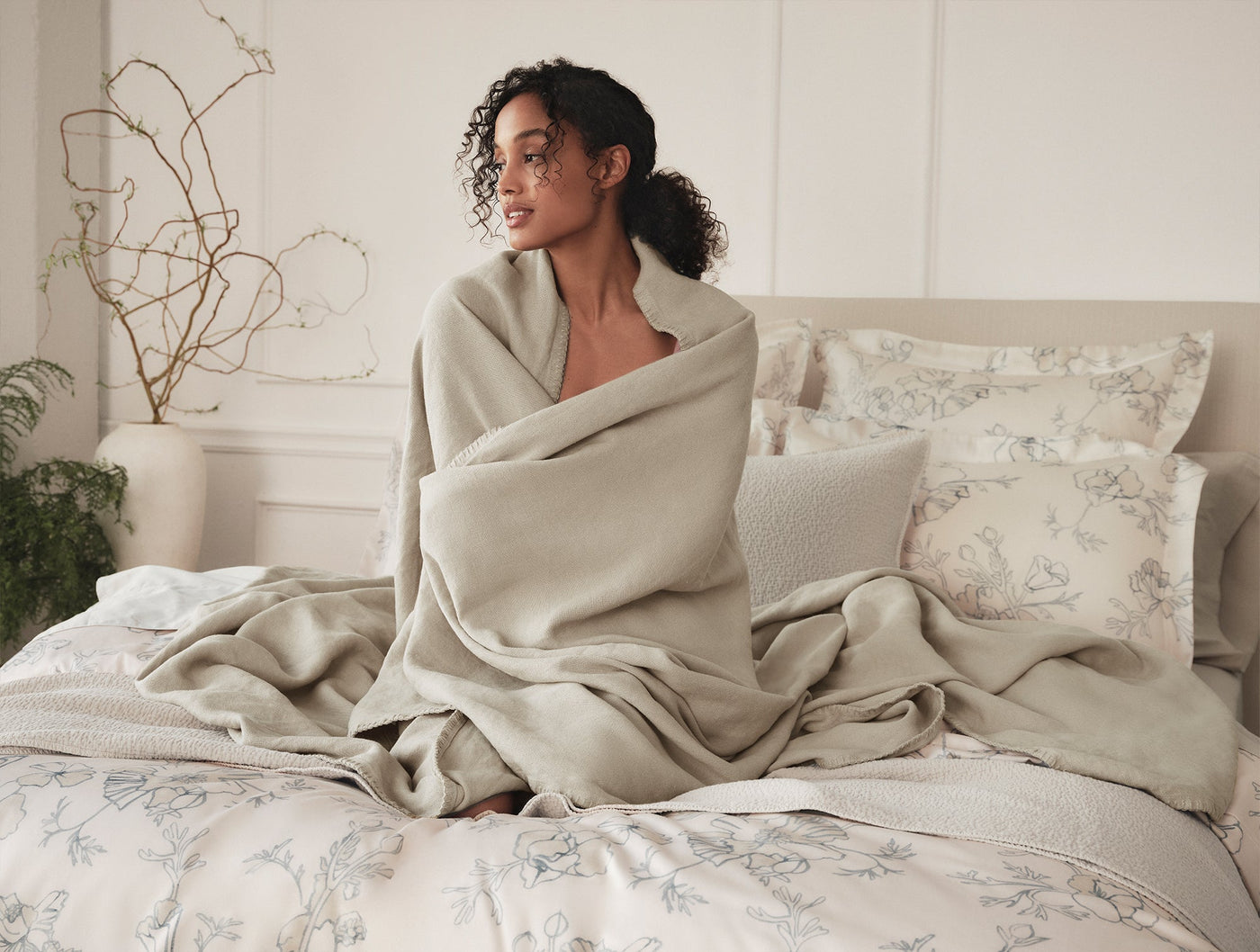 Woman wrapped in a sage blanket seated on a bed styled with Romneya Print shams.