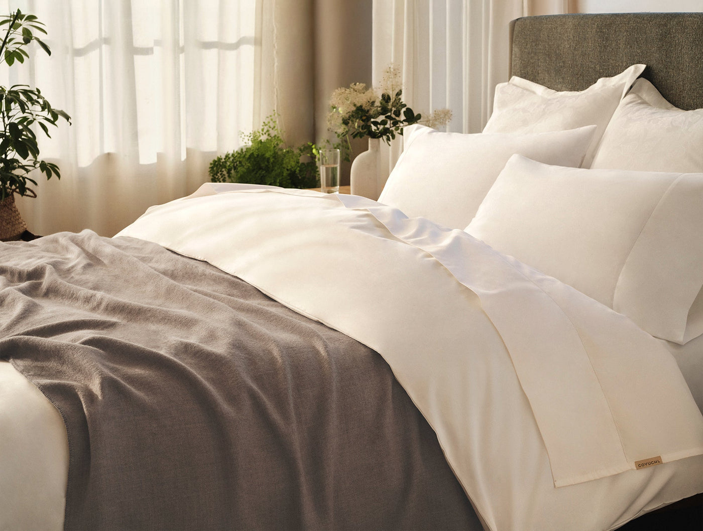 Ivory sateen sheet set styled on a bed with a taupe throw blanket and sheer window backdrop.