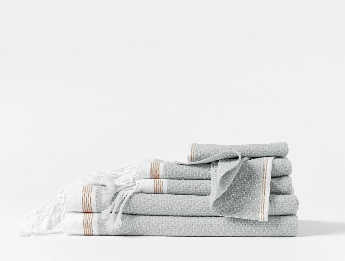 Stacked mist grey waffle-weave towels with tan stripe trim and fringed edge detail.