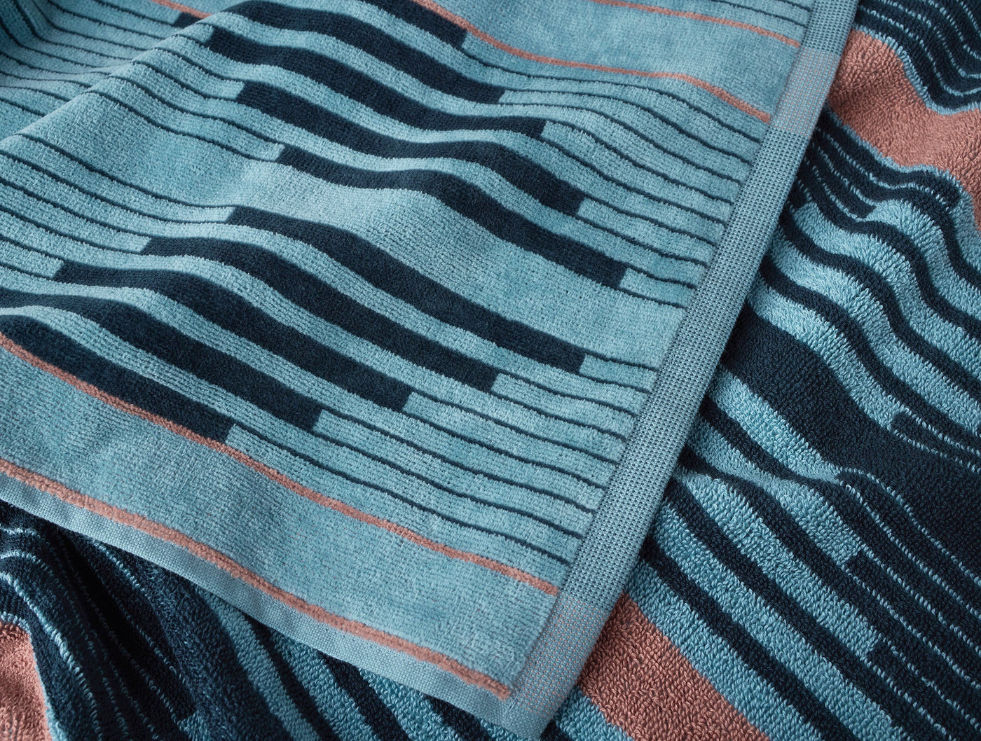 Close-up of Coyuchi Morelia beach towel showing teal wavy stripe pattern and rust hem detail.