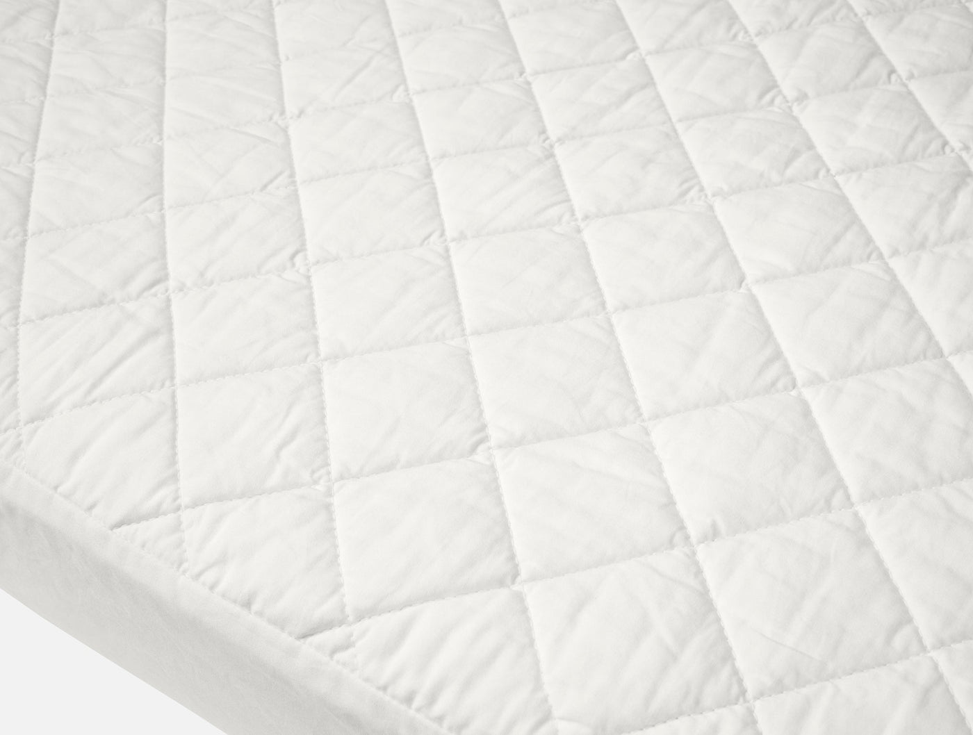 Close-up of Coyuchi ivory quilted cotton mattress pad showing diamond stitch pattern and fitted skirt.