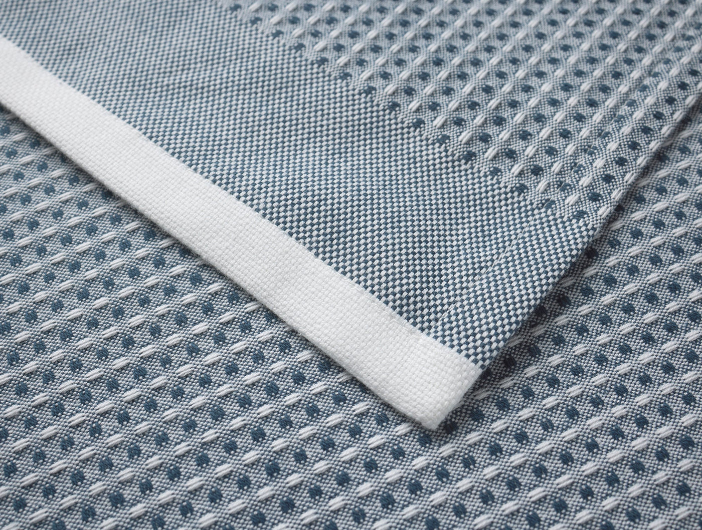 Close-up of Coyuchi Waffle kitchen towel corner showing white hem detail on slate blue waffle-weave fabric.